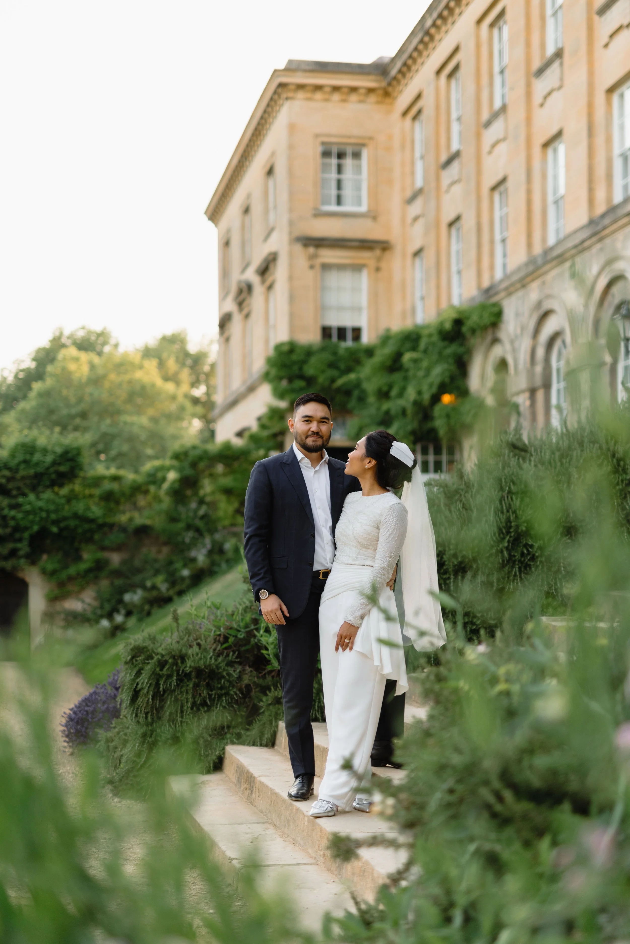 A wedding couple standing on a stone pathway surrounded by greenery, with a historic building in the background.