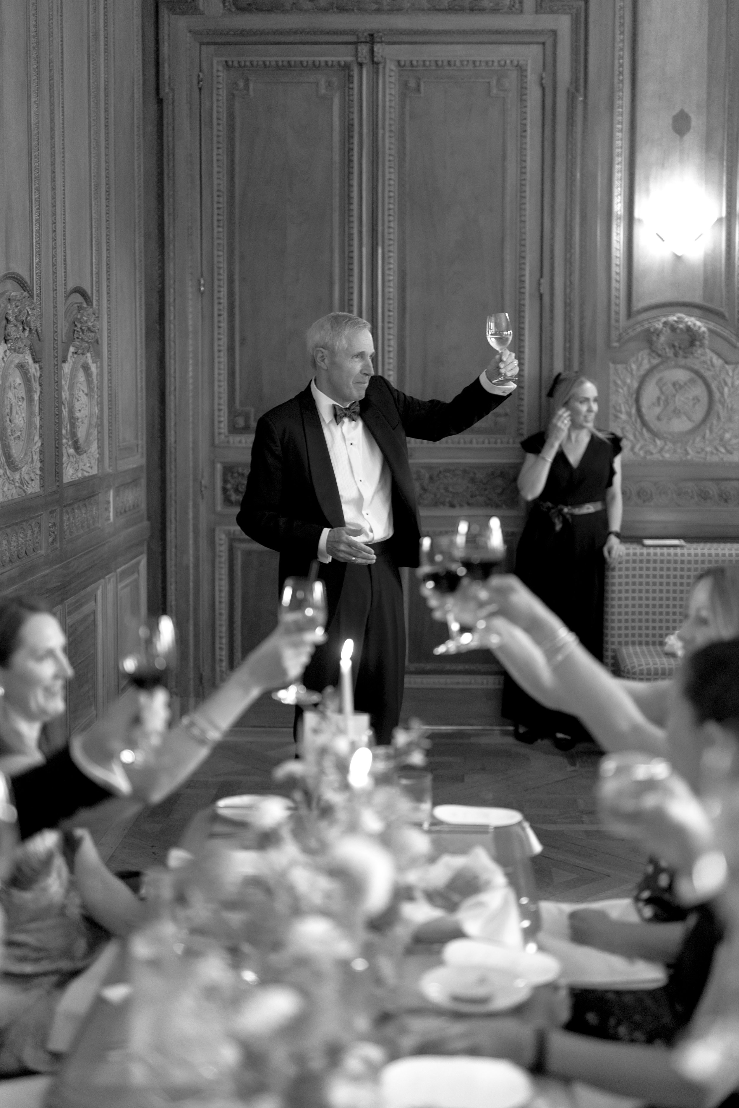 An elderly man in a tuxedo raising a glass at a formal dinner celebration. Women are seated at a table, some also holding glasses, with a decorated table setting including flowers and candles. The setting appears to be an elegant, wood-paneled room.
