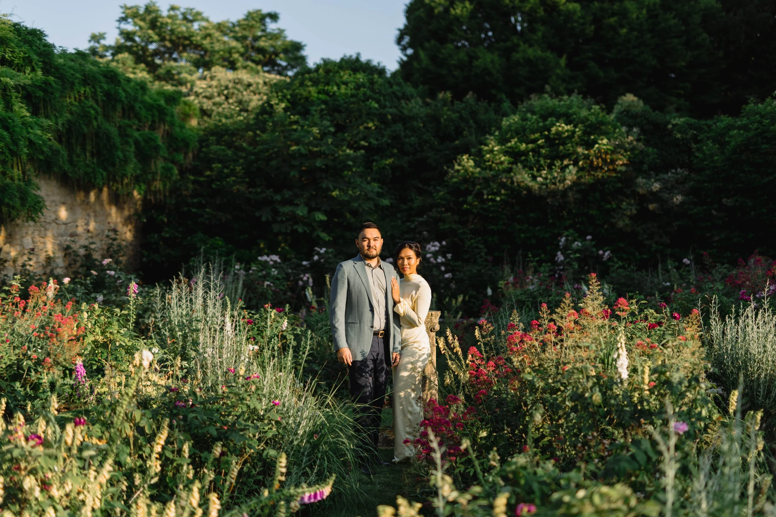 A couple standing in a lush garden with various flowers and greenery, with trees in the background. The man is wearing a gray suit and the woman is in a light-colored dress.