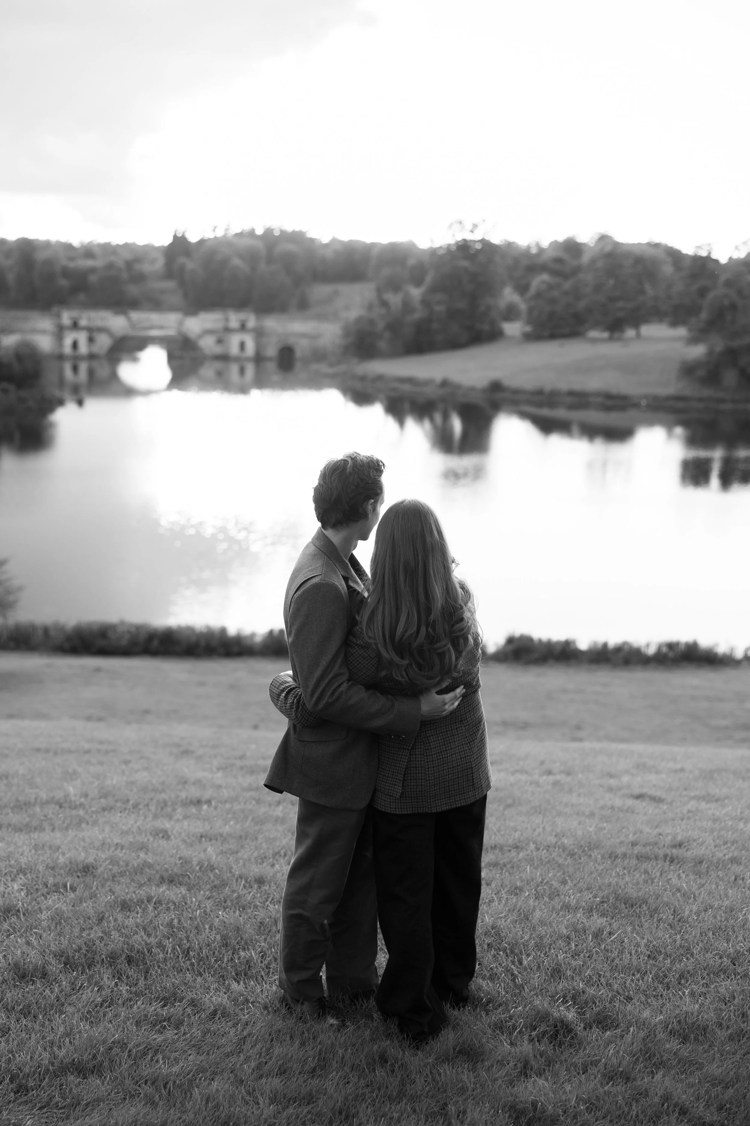 A black-and-white photo of a couple embracing by a lake, with trees and a bridge in the background.