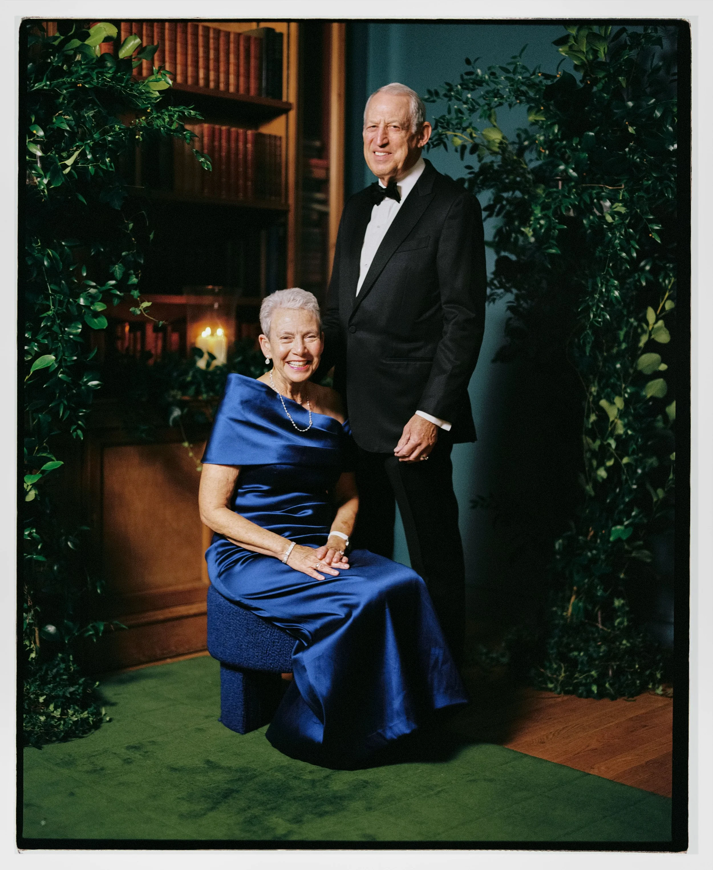 An elder woman in a blue gown sitting on a stool and smiling, accompanied by a man in a black tuxedo standing beside her in a warmly lit room with bookshelves, green plants, and a candle in the background.