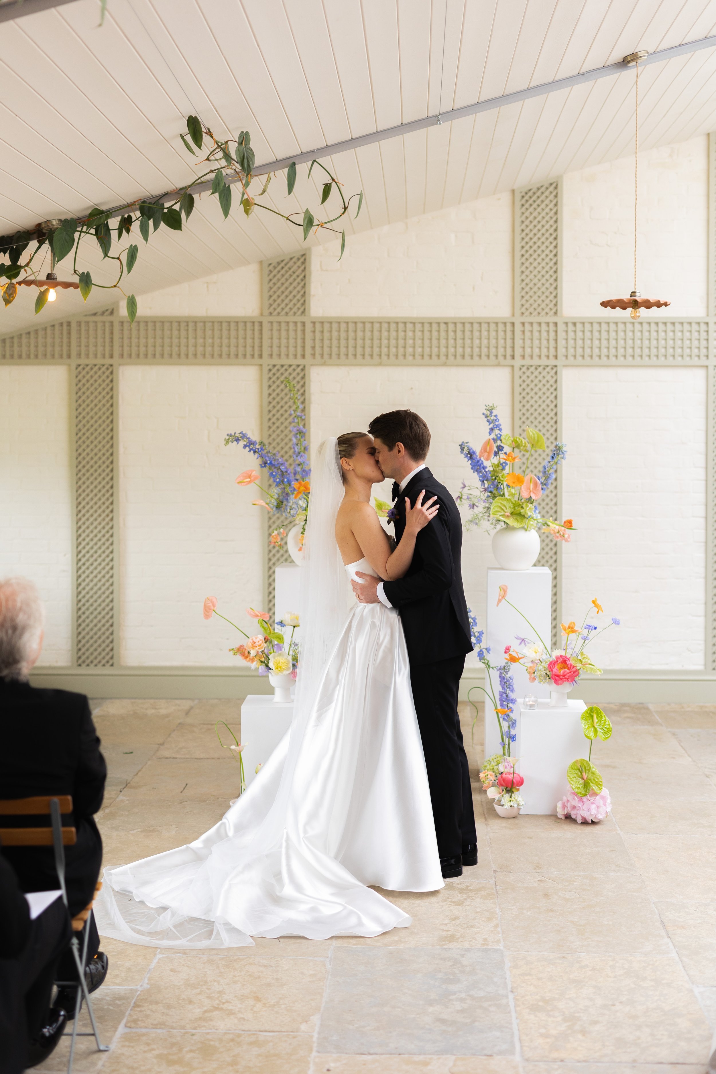 A bride and groom sharing a kiss during their wedding ceremony, with guests and floral arrangements visible in the background.