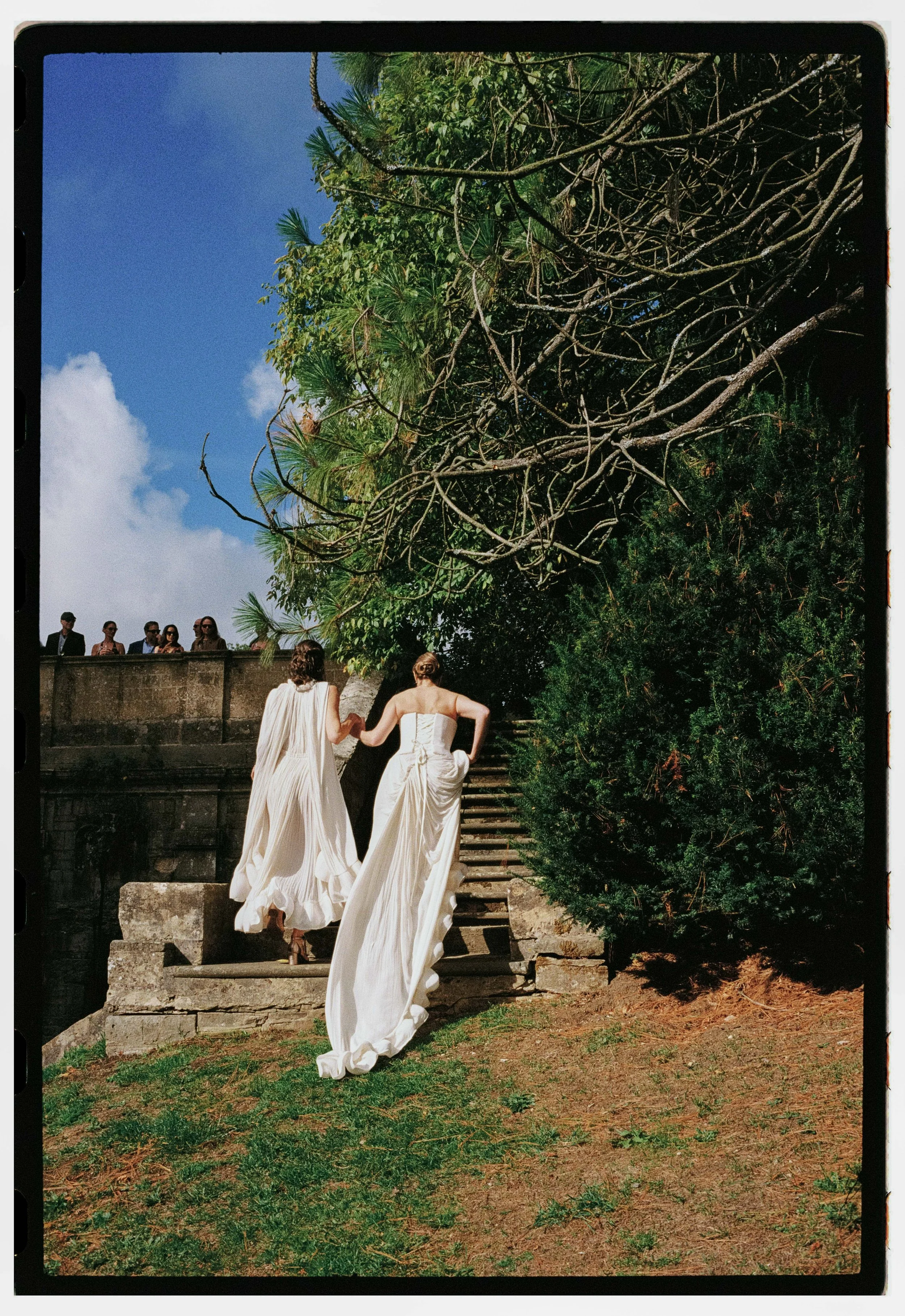 Two brides in white dresses climbing stone steps outdoors, with onlookers watching from a stone wall, greenery, trees, and a blue sky in the background.
