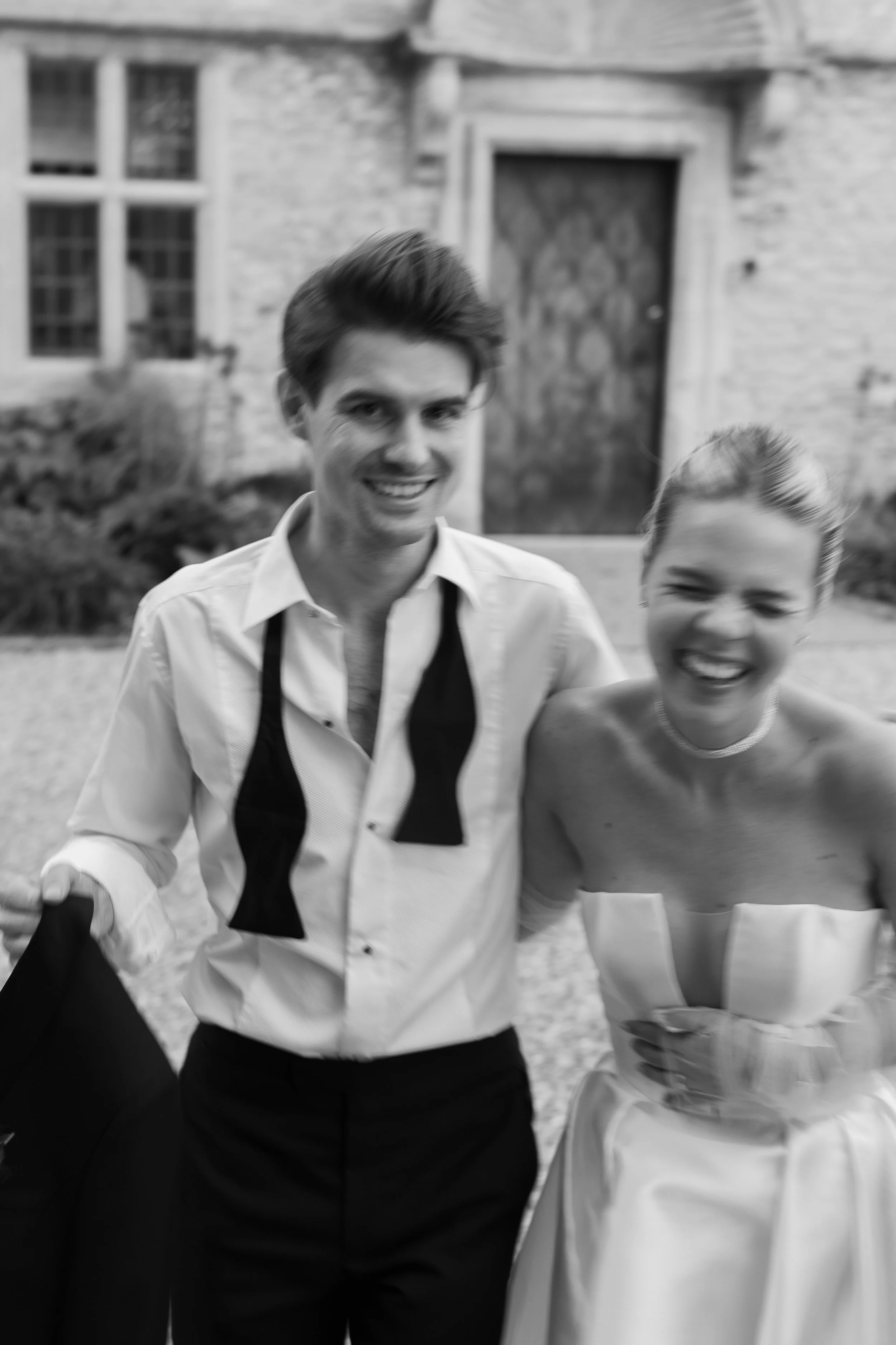 Black and white photo of a young man and woman smiling and standing outside in front of a brick building. The man has styled hair and is wearing a shirt with a black bow tie, which is untied and hanging around his neck. The woman is wearing a straple