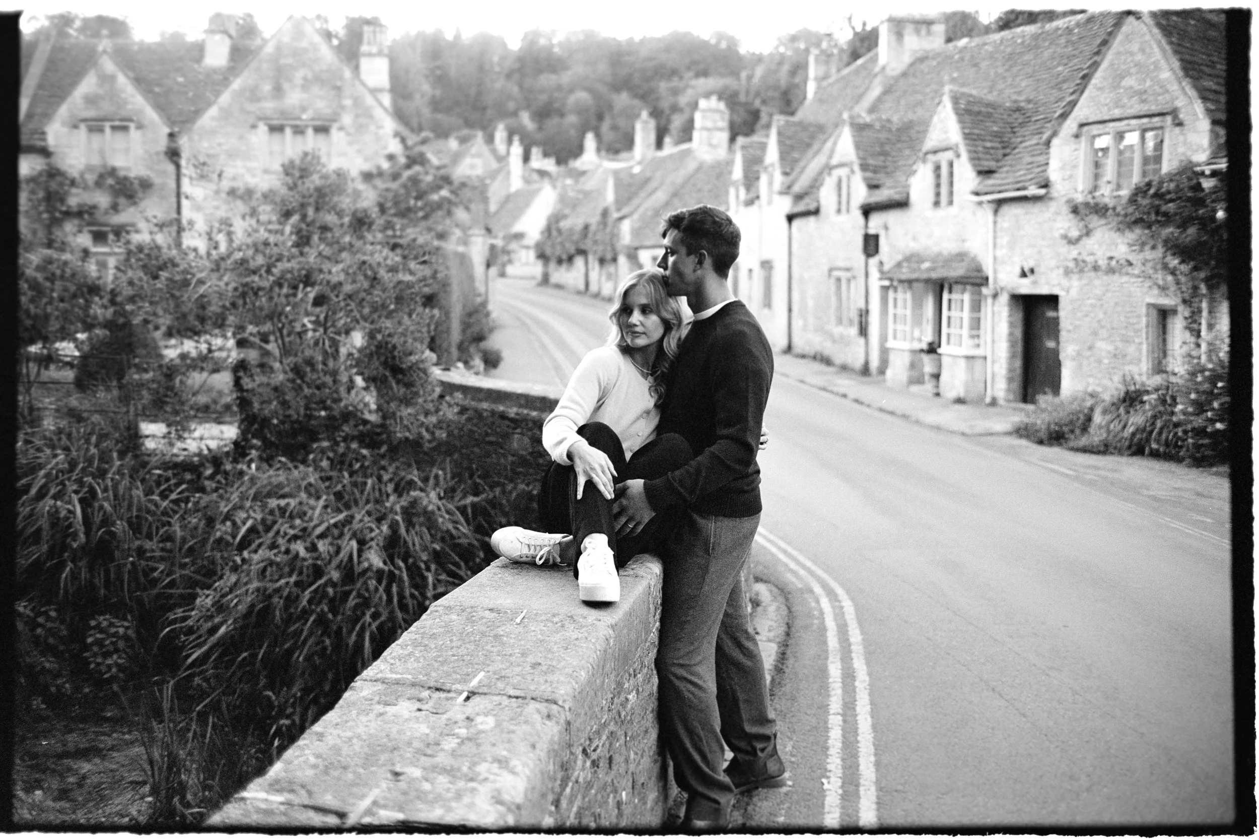 A young couple sitting on a stone wall on a quiet street in a small town, with houses and trees in the background.