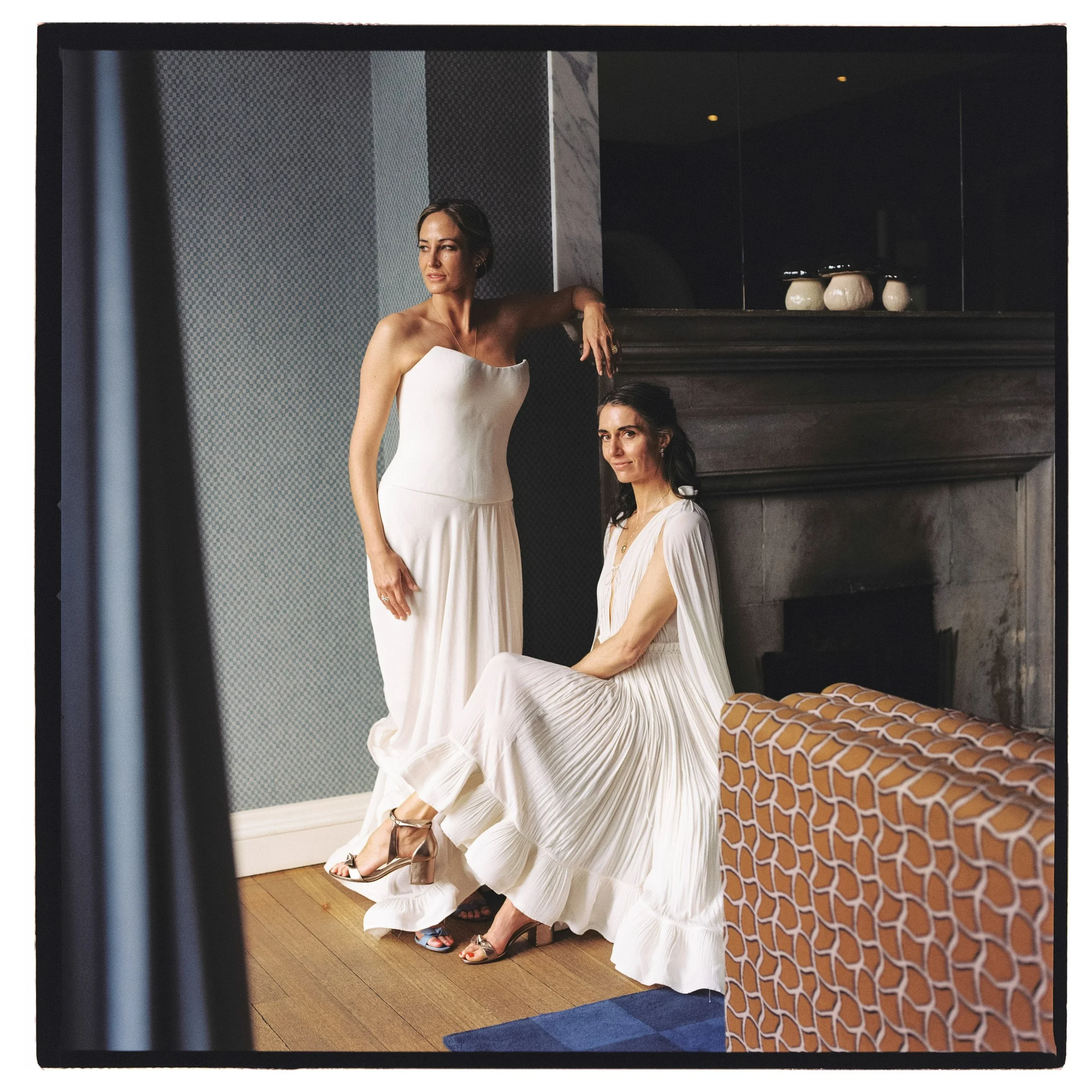 Two brides in white wedding dresses, one standing and one sitting, in a room with a fireplace and patterned orange and blue furniture.