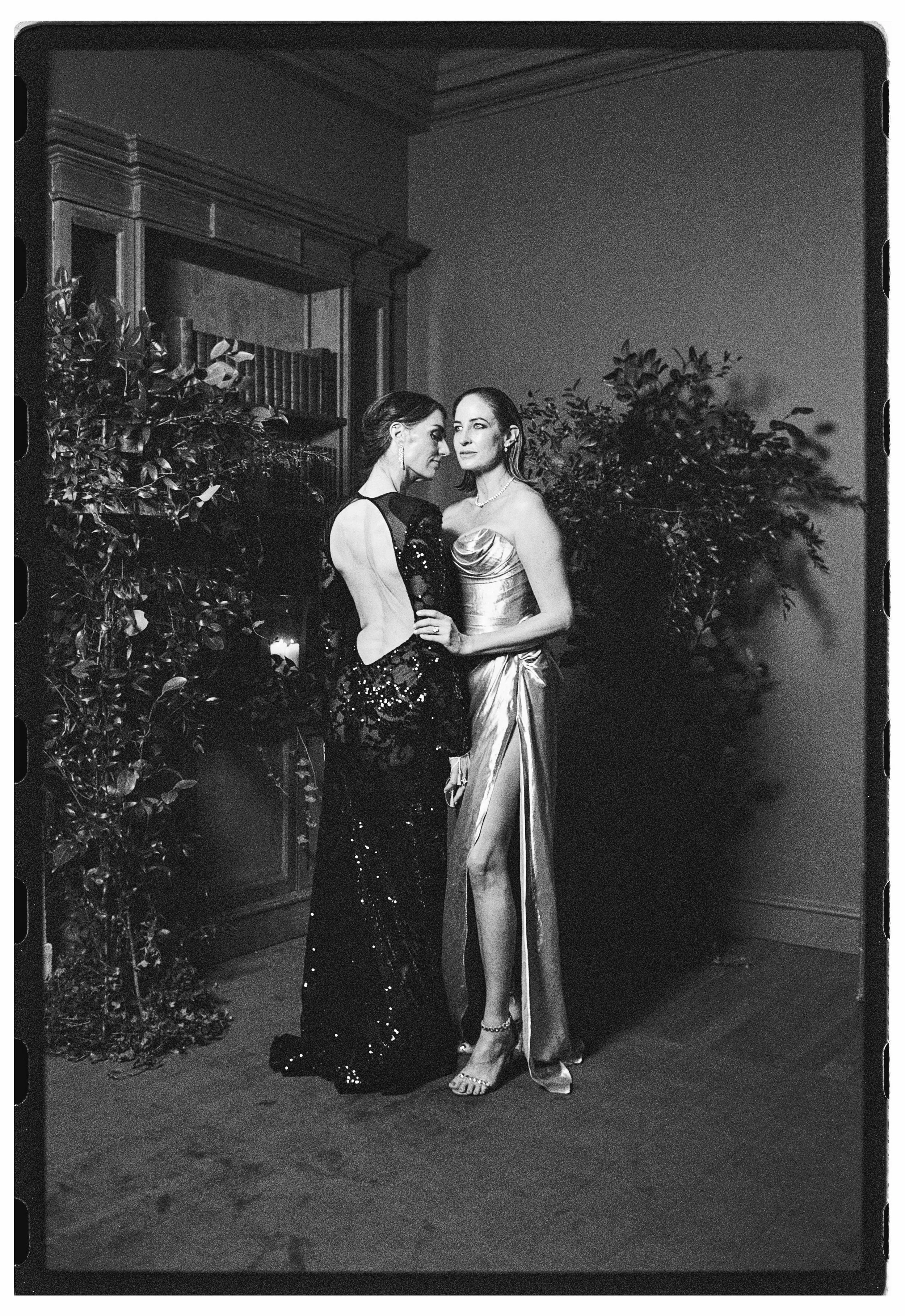 The brides in elegant gowns stand close together in a formal setting, surrounded by plants and a wooden cabinet.