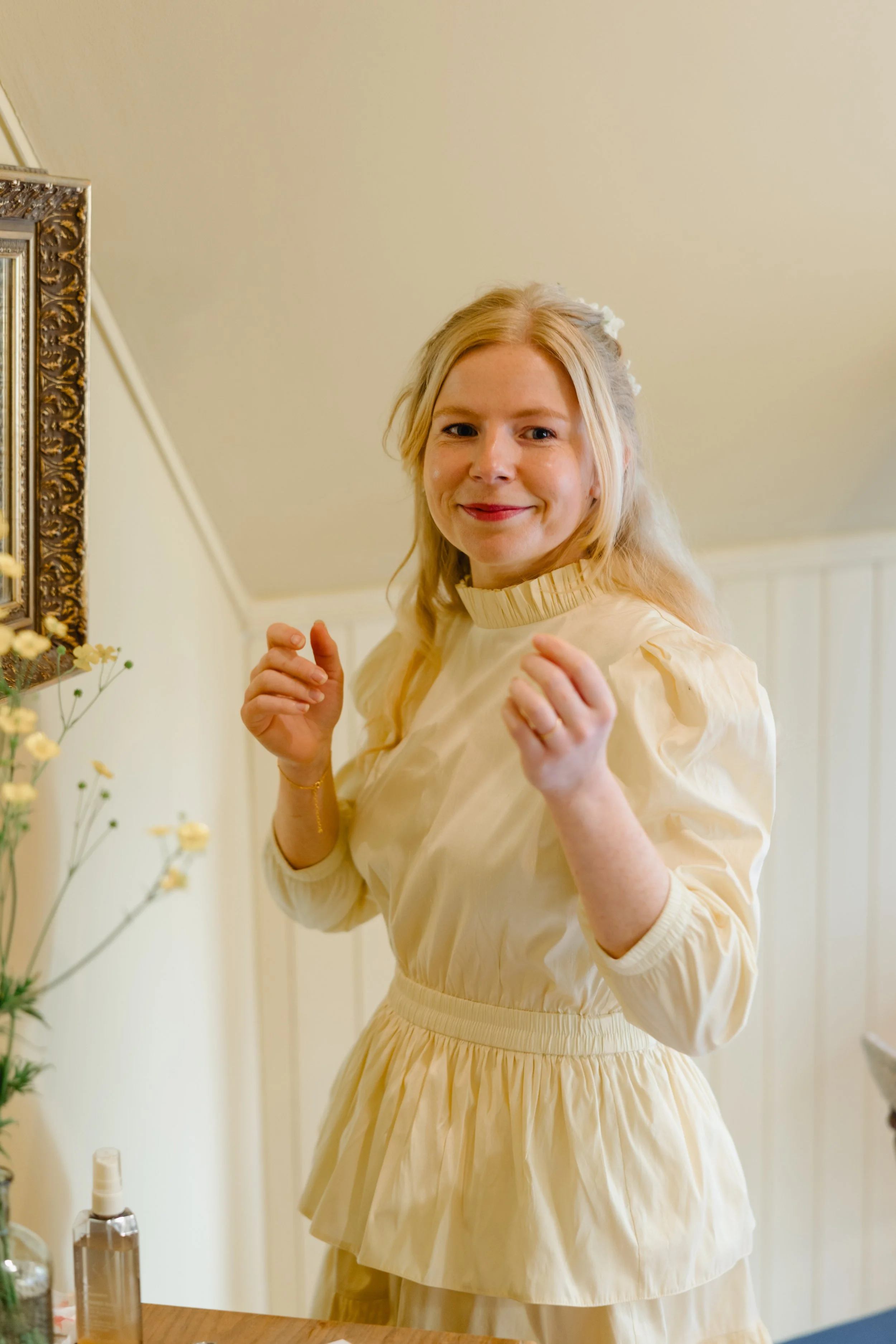 A smiling woman in a cream-colored dress with puffy sleeves and a ruffled collar standing in a room with cream-colored walls and a framed mirror.