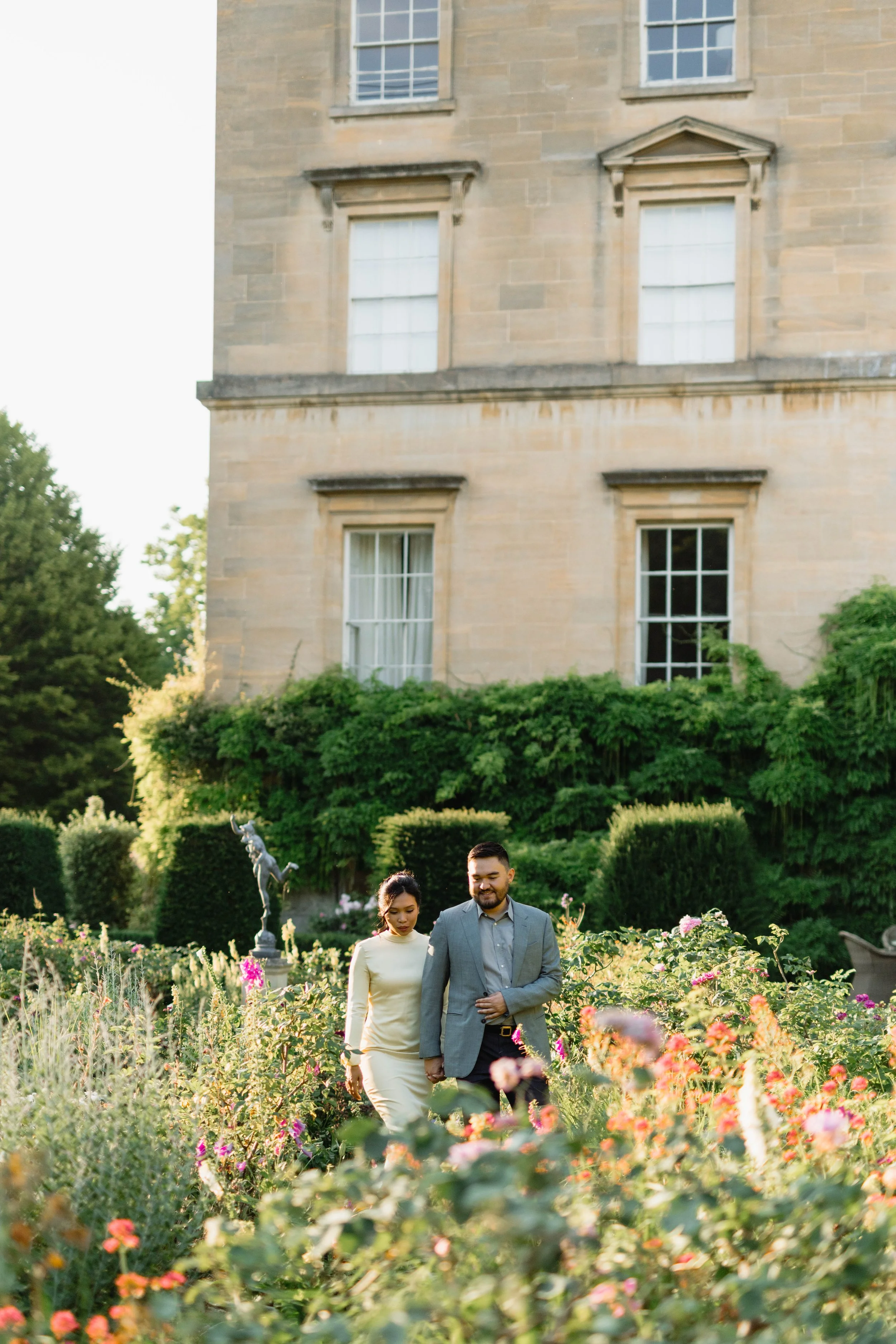 A man and woman walking in a garden full of flowers, with a large stone building in the background.
