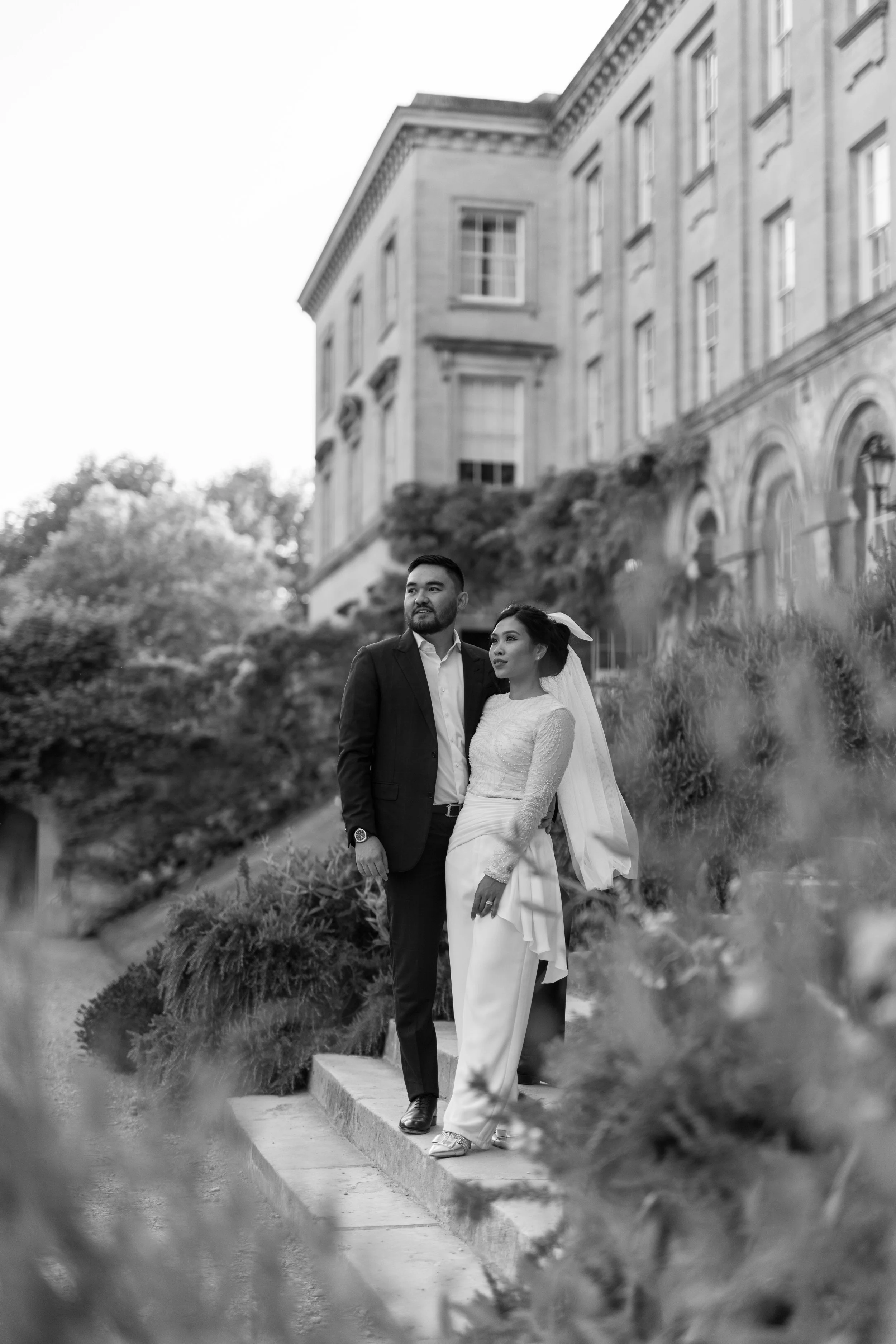 Black and white photo of a couple standing outdoors, with a large historic building and trees in the background. The man is wearing a suit, and the woman is dressed in elegant attire with a veil.