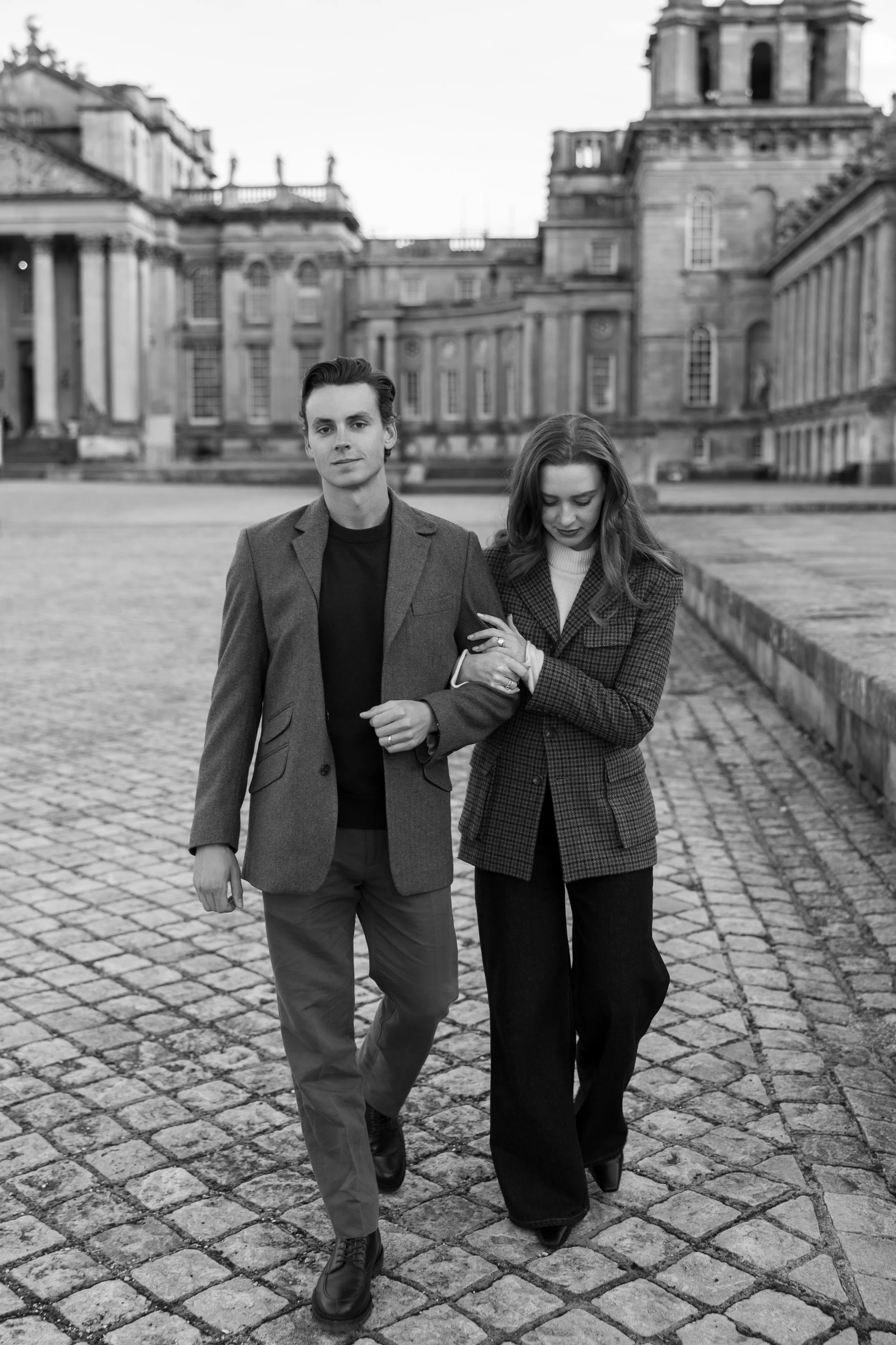 A black and white photo of a man and woman walking arm in arm on a cobblestone street with historic buildings in the background.