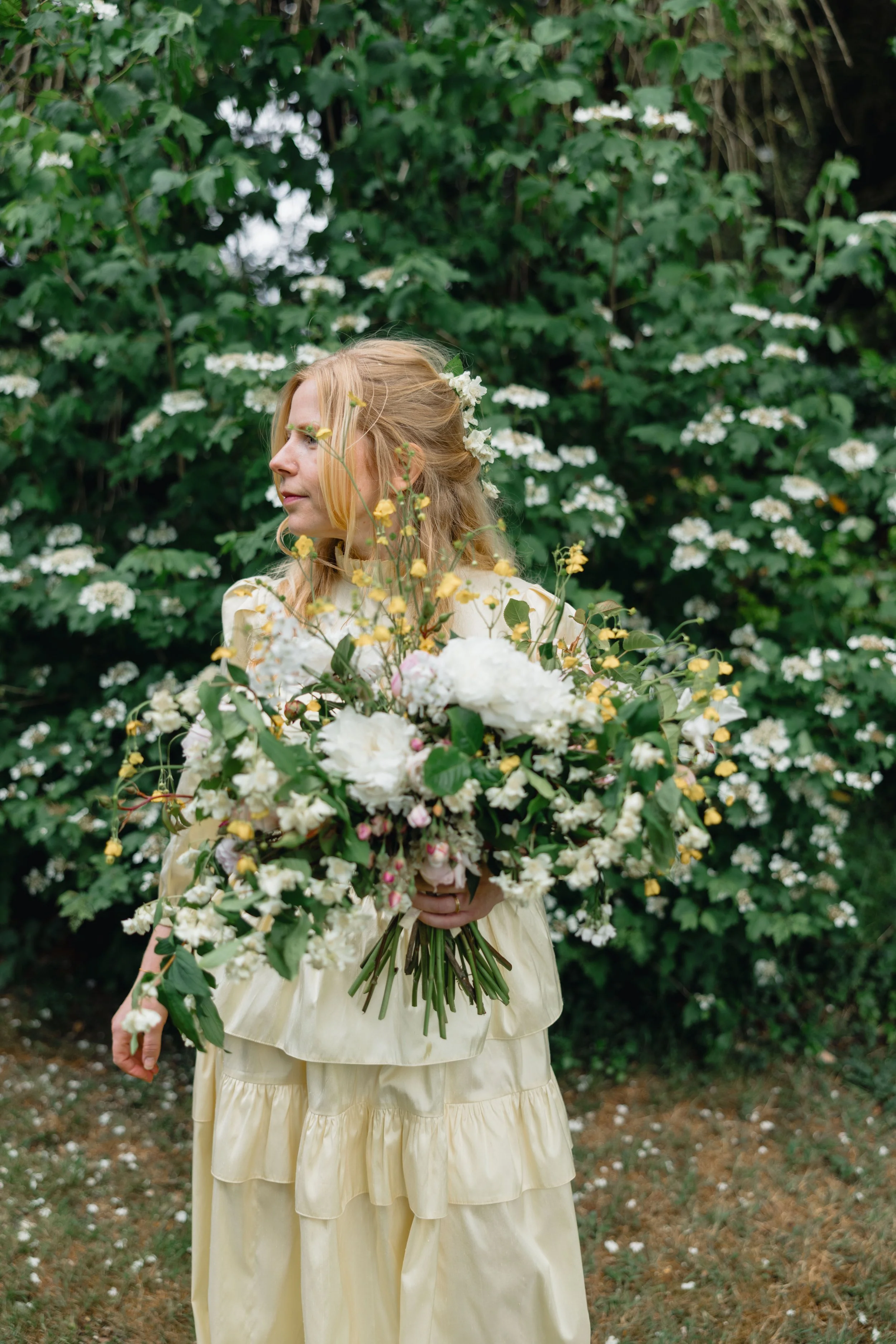 Woman with long, red hair decorated with small white flowers, holding a large bouquet of white, pink, and yellow flowers, standing outdoors in front of green foliage and white flowering bushes.