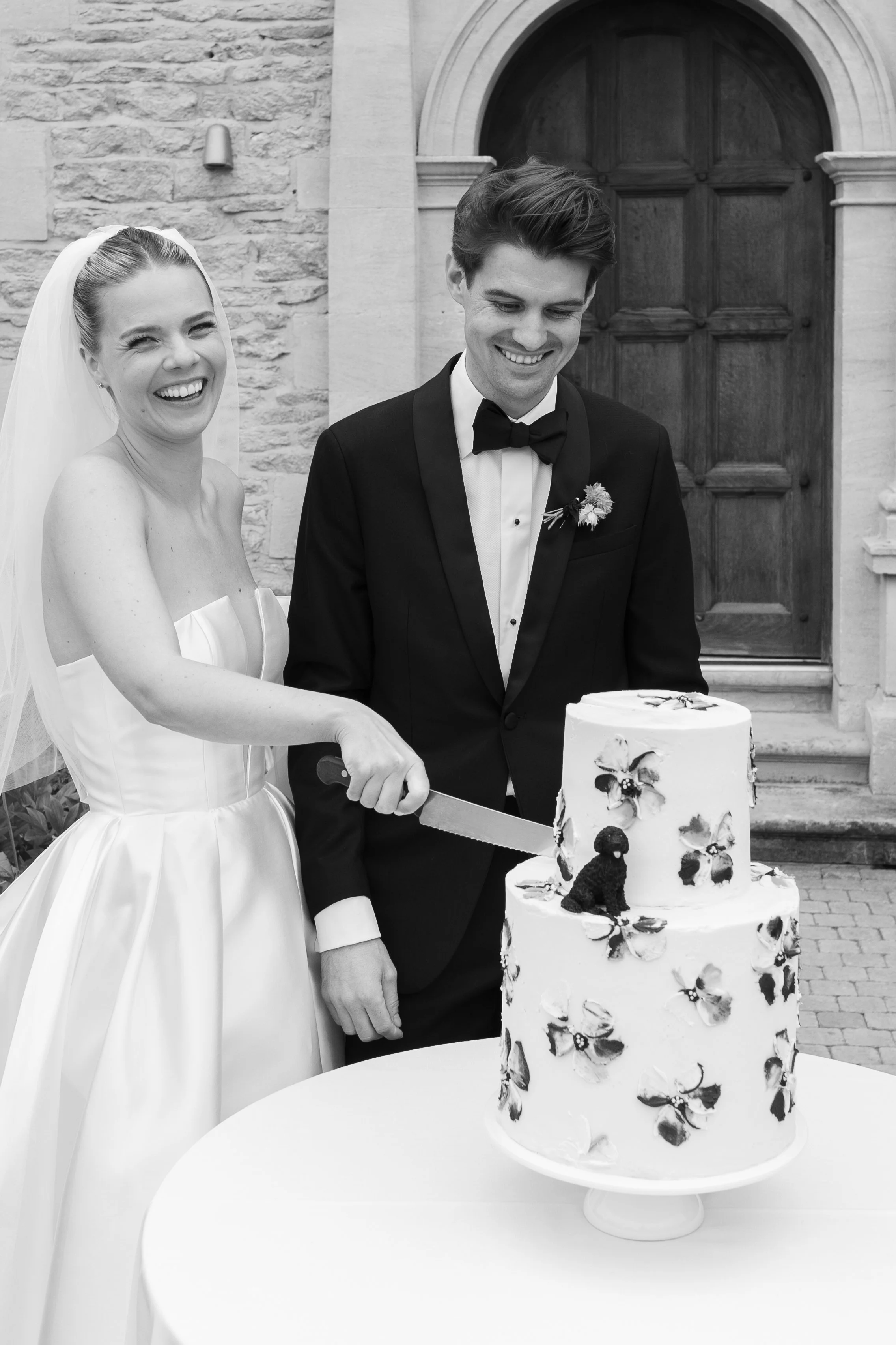 A bride and groom smiling as they cut a wedding cake outdoors. The cake has two tiers decorated with floral motifs and a small dog figurine on top.