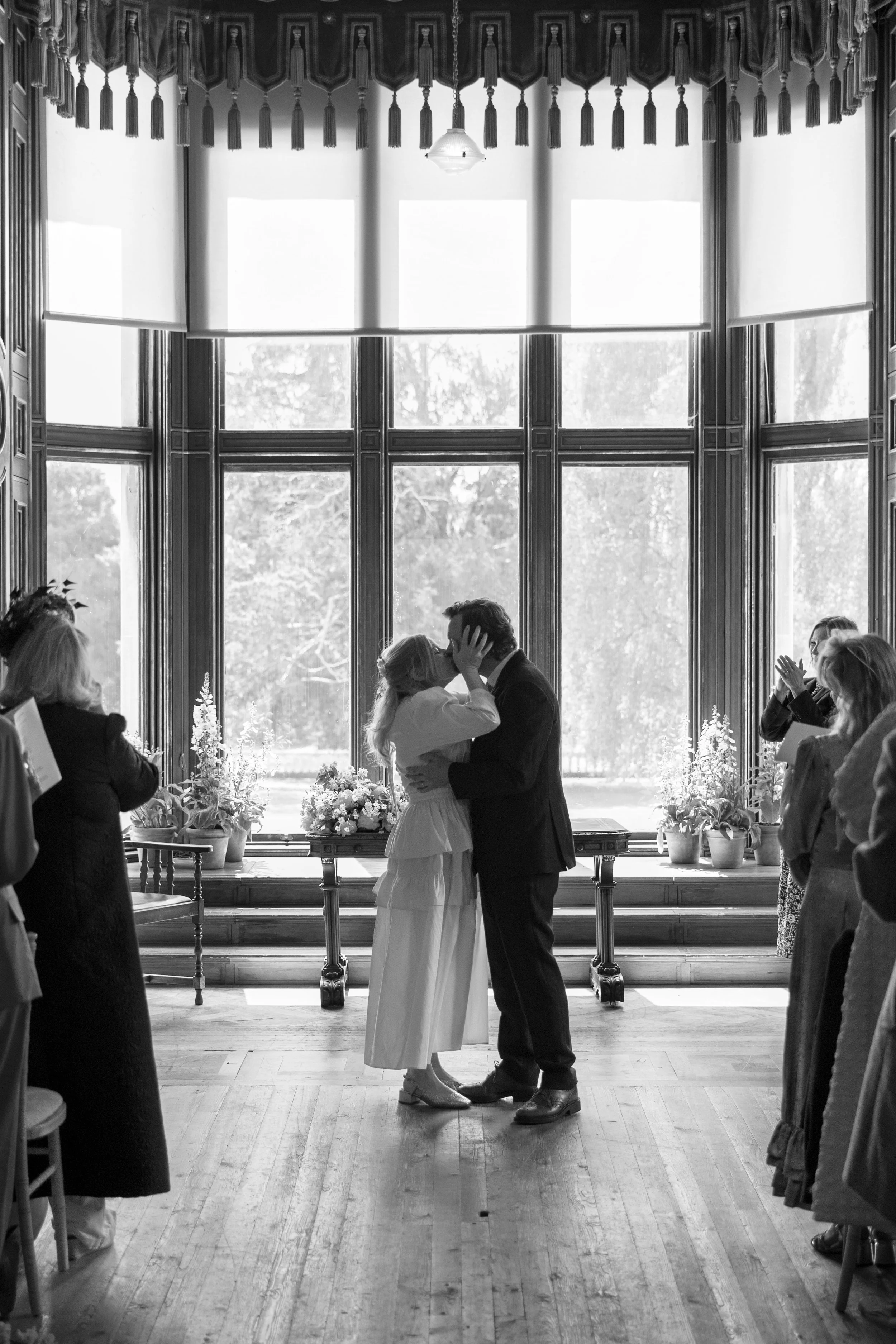 A black and white photo of a couple sharing a kiss during their wedding ceremony in front of a large window with tall curtains. Wedding guests are clapping and watching nearby.