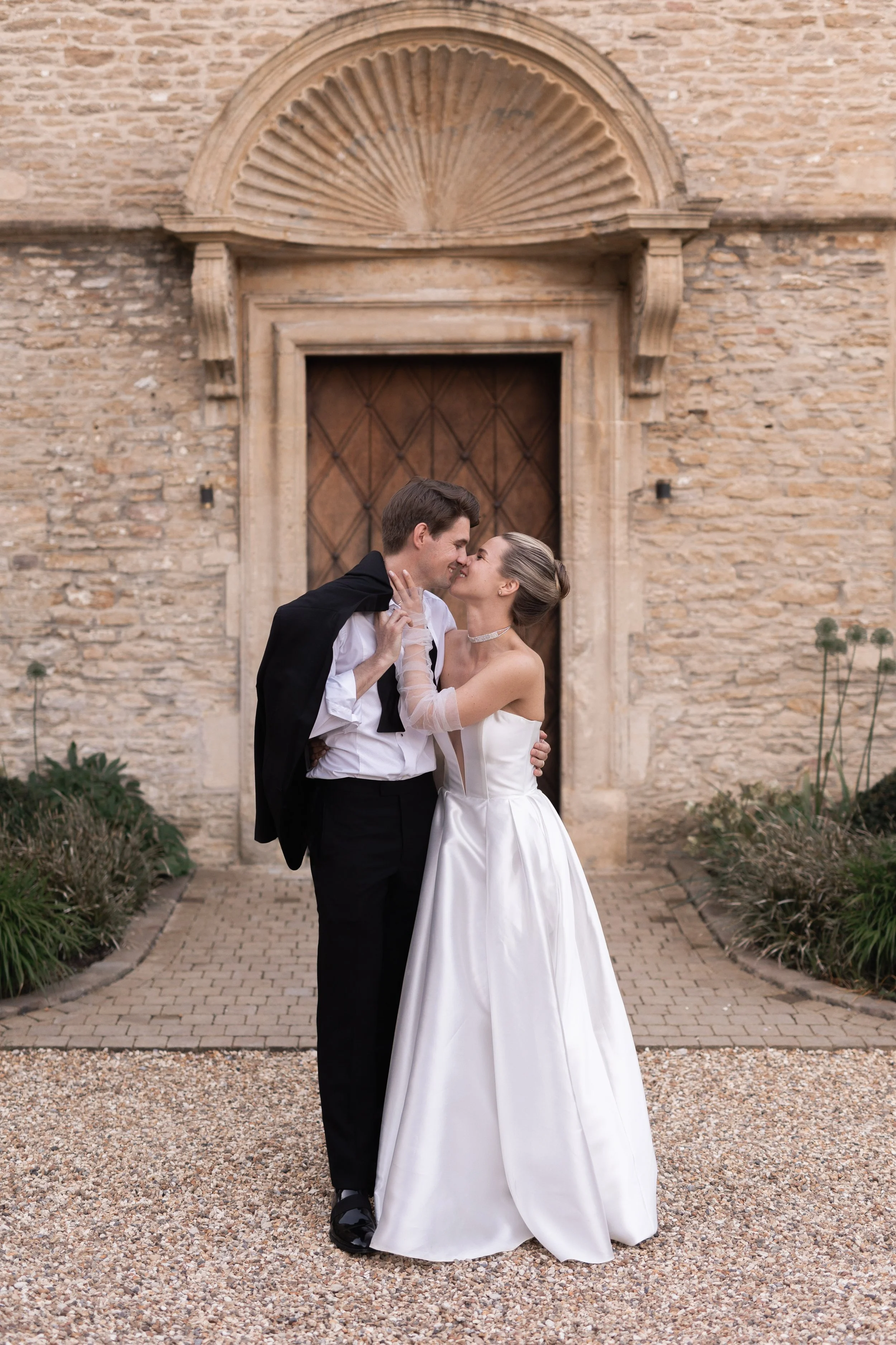 A newlywed couple sharing a kiss in front of a historic stone building with a wooden door and an ornate stone archway.