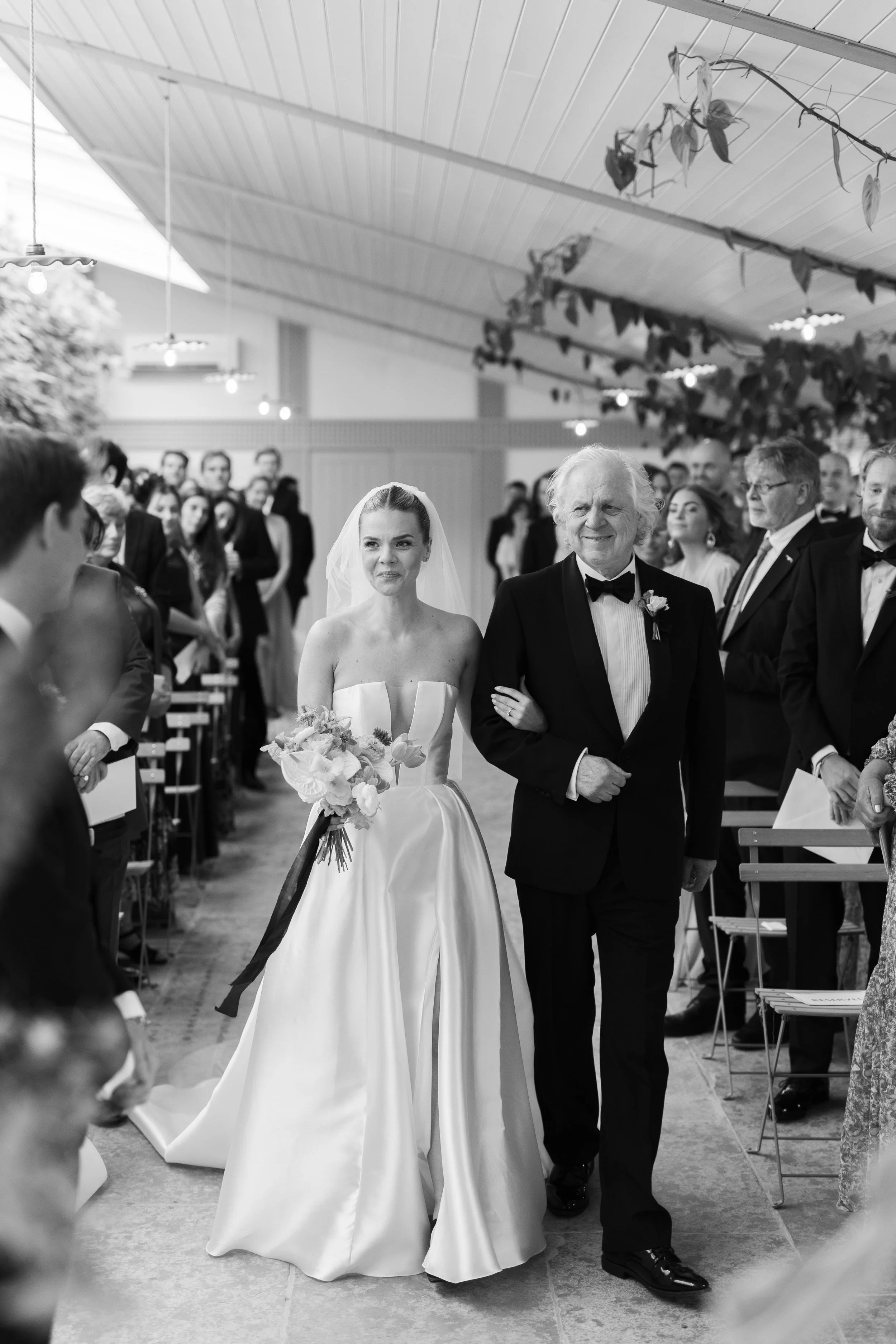 A bride in a strapless wedding gown holding a bouquet walking down the aisle with an older man in a tuxedo at a wedding ceremony inside a decorated venue.