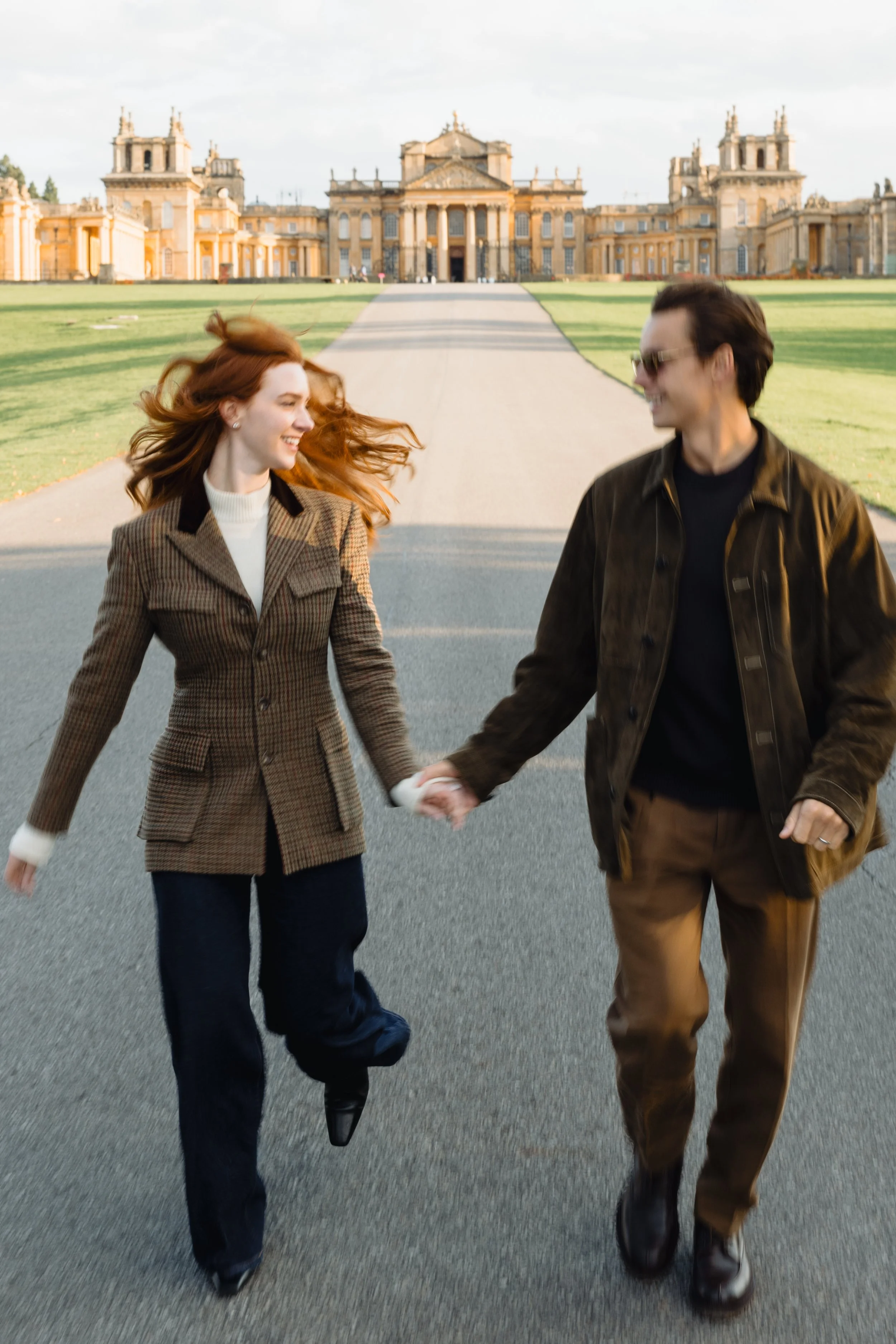 A couple holding hands and walking together outdoors on a paved road with a grand historic building in the background. The woman has red hair and is smiling, while the man wears sunglasses and looks at her.