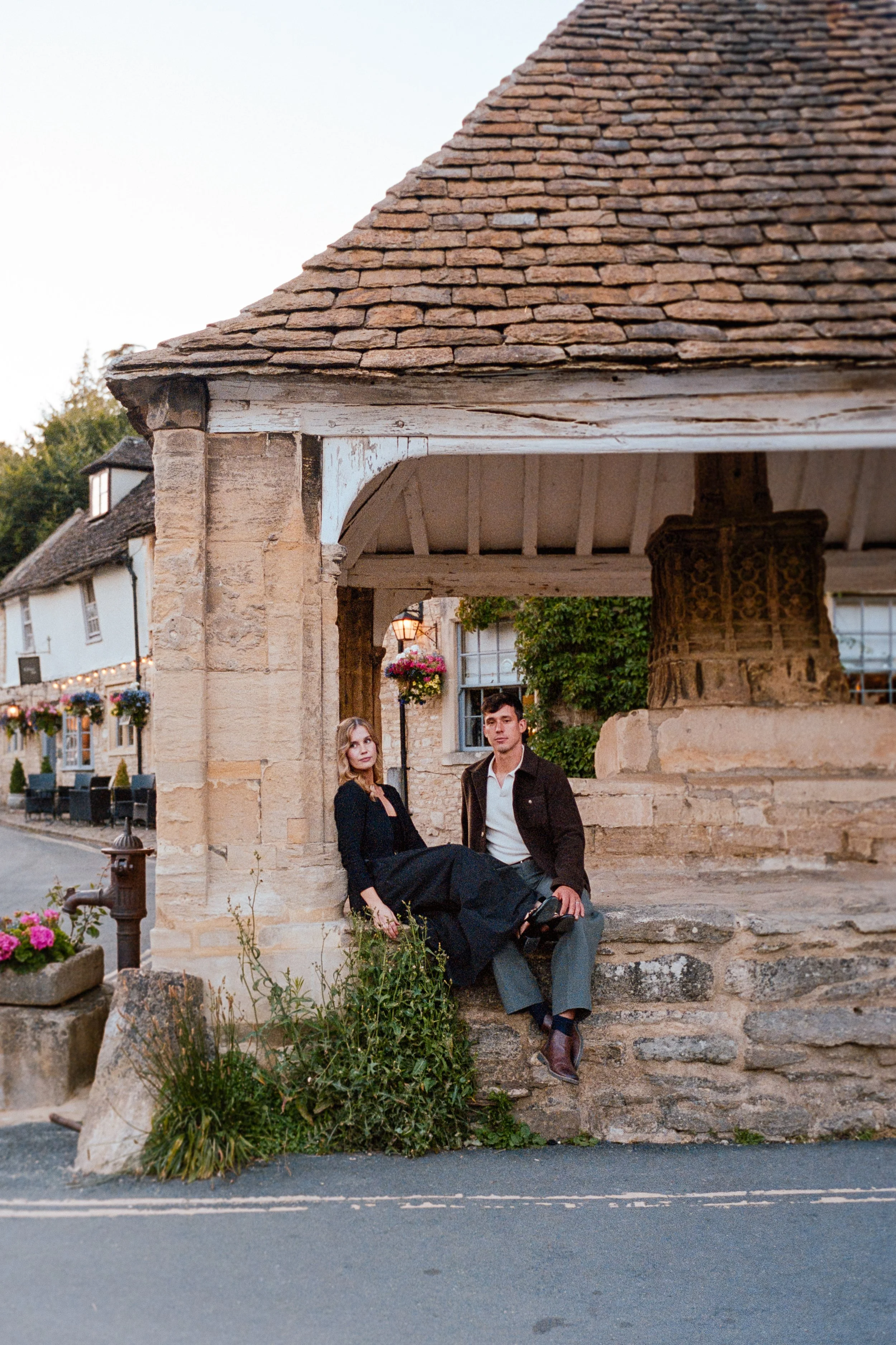 A man and woman sitting on stone steps under a historical stone well with a wooden roof, in a small village street with hanging flower baskets and stone buildings.