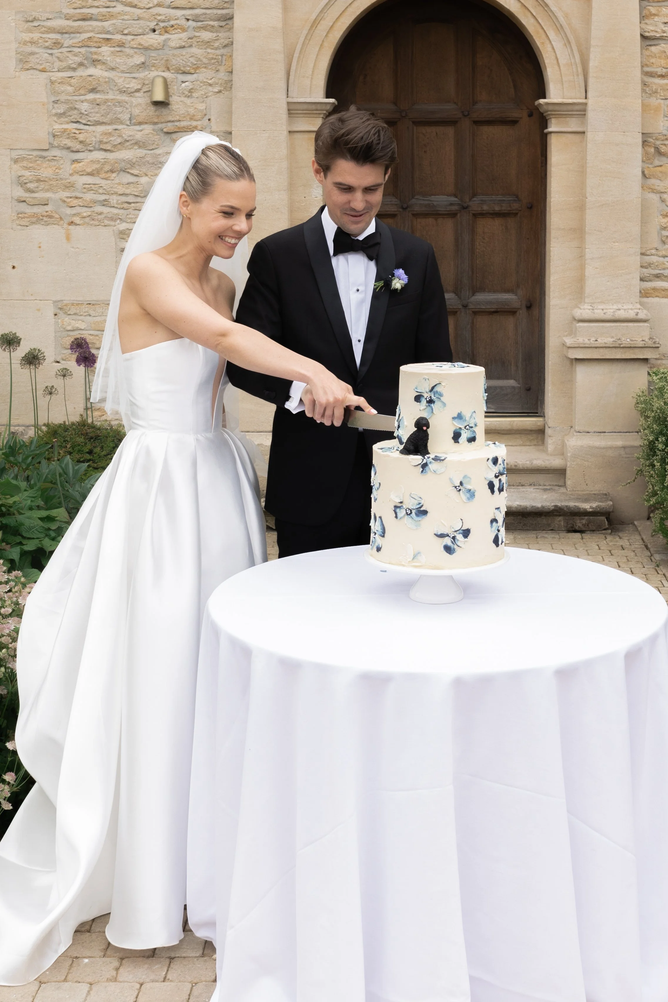 A bride and groom in wedding attire are cutting a tiered wedding cake decorated with blue and white flowers outside a stone building.