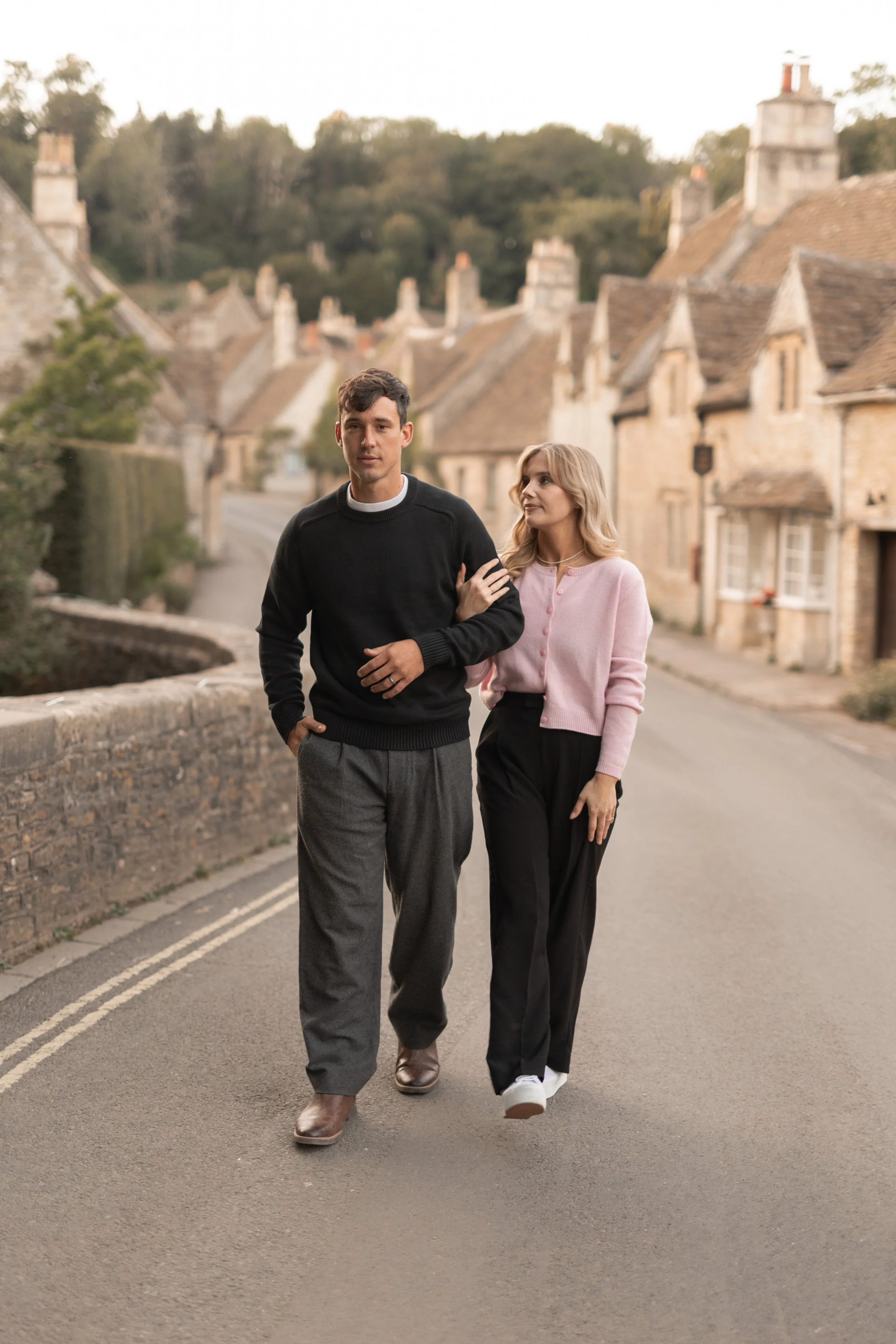 A young man and woman walking arm in arm down a quiet, stone-lined street in a village with beige stone cottages with tiled roofs, surrounded by greenery.