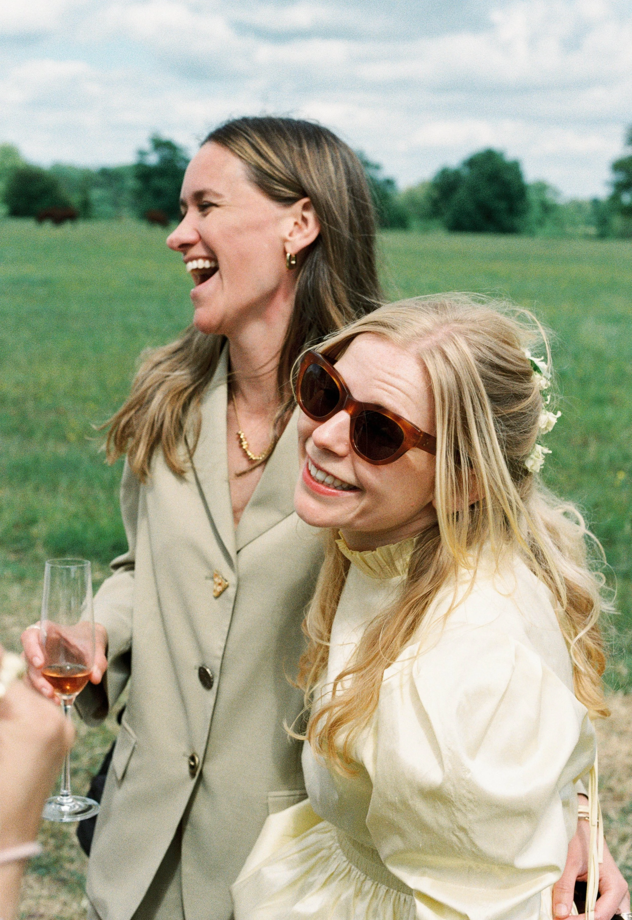 Two women laughing outdoors in a field, one with sunglasses and the other with glasses, holding a glass of wine.