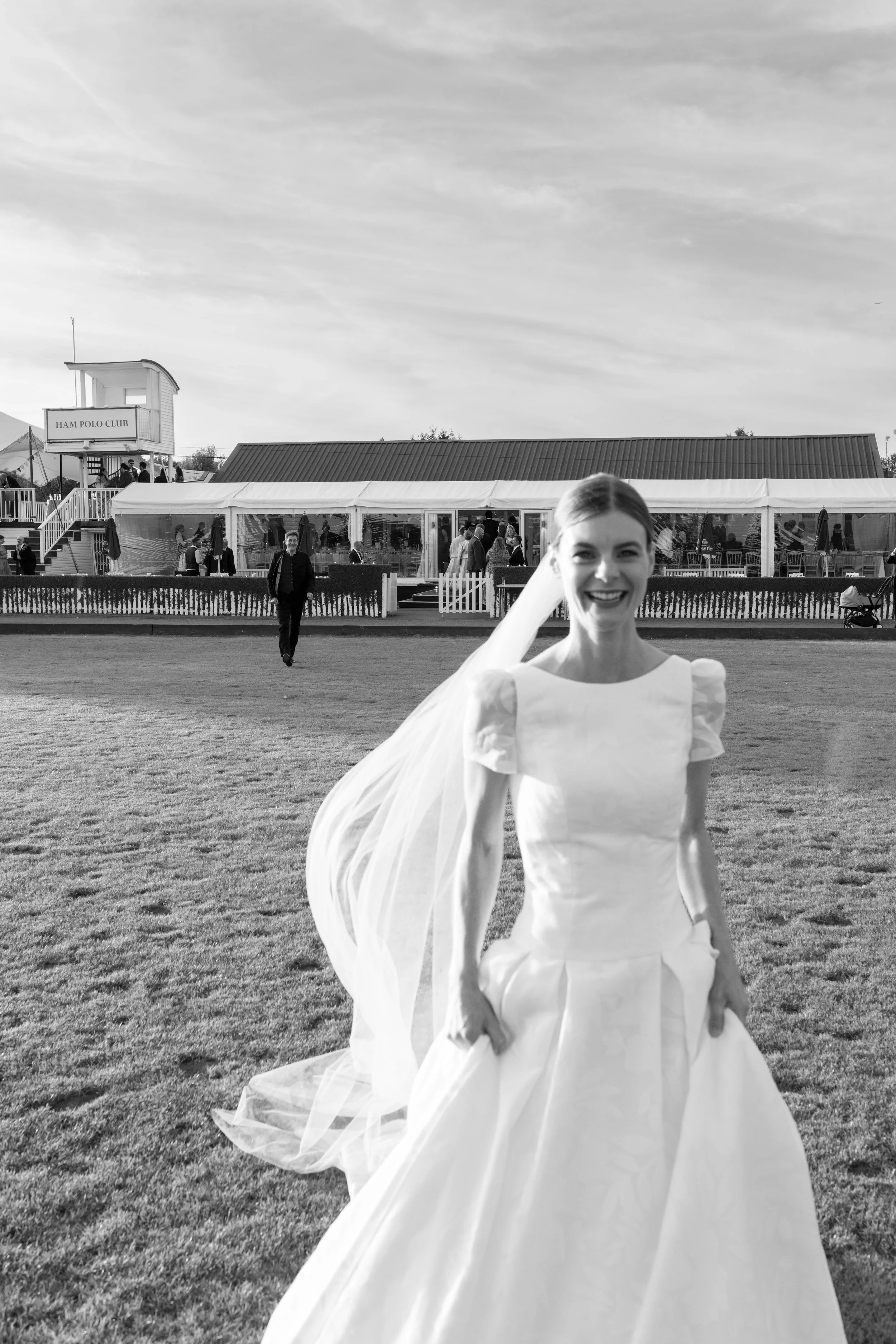 A smiling woman in a wedding dress standing outdoors with a large tent and Ham Polo Club in the background.