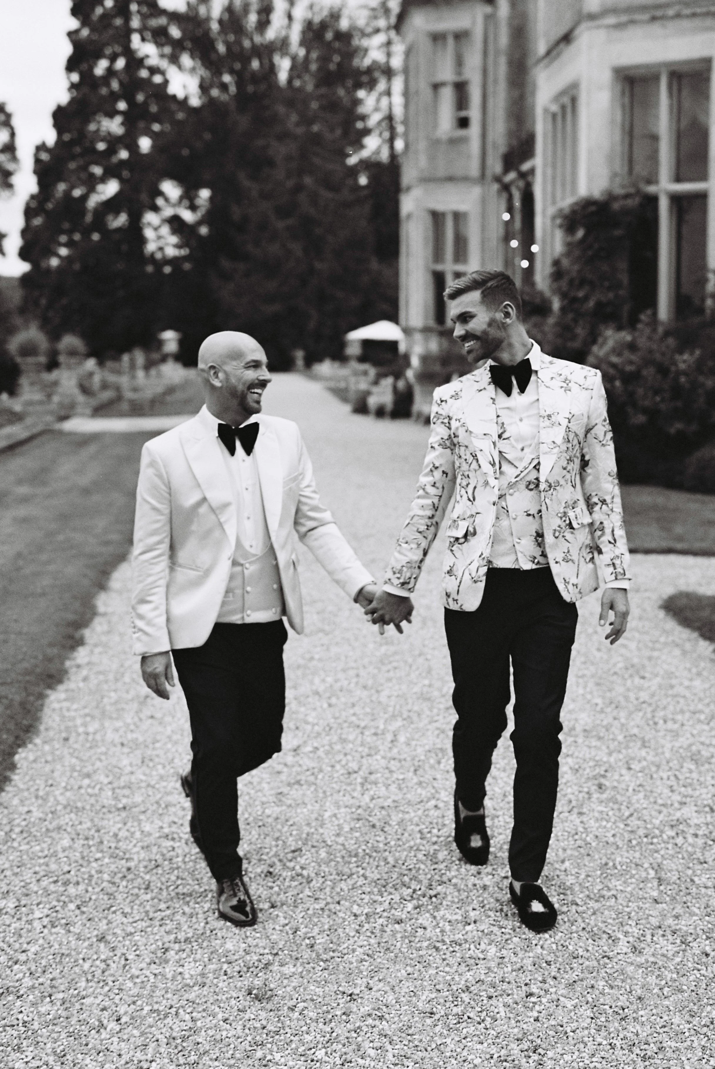 Two men in tuxedos holding hands and smiling while walking on a gravel driveway outside a large estate or mansion.