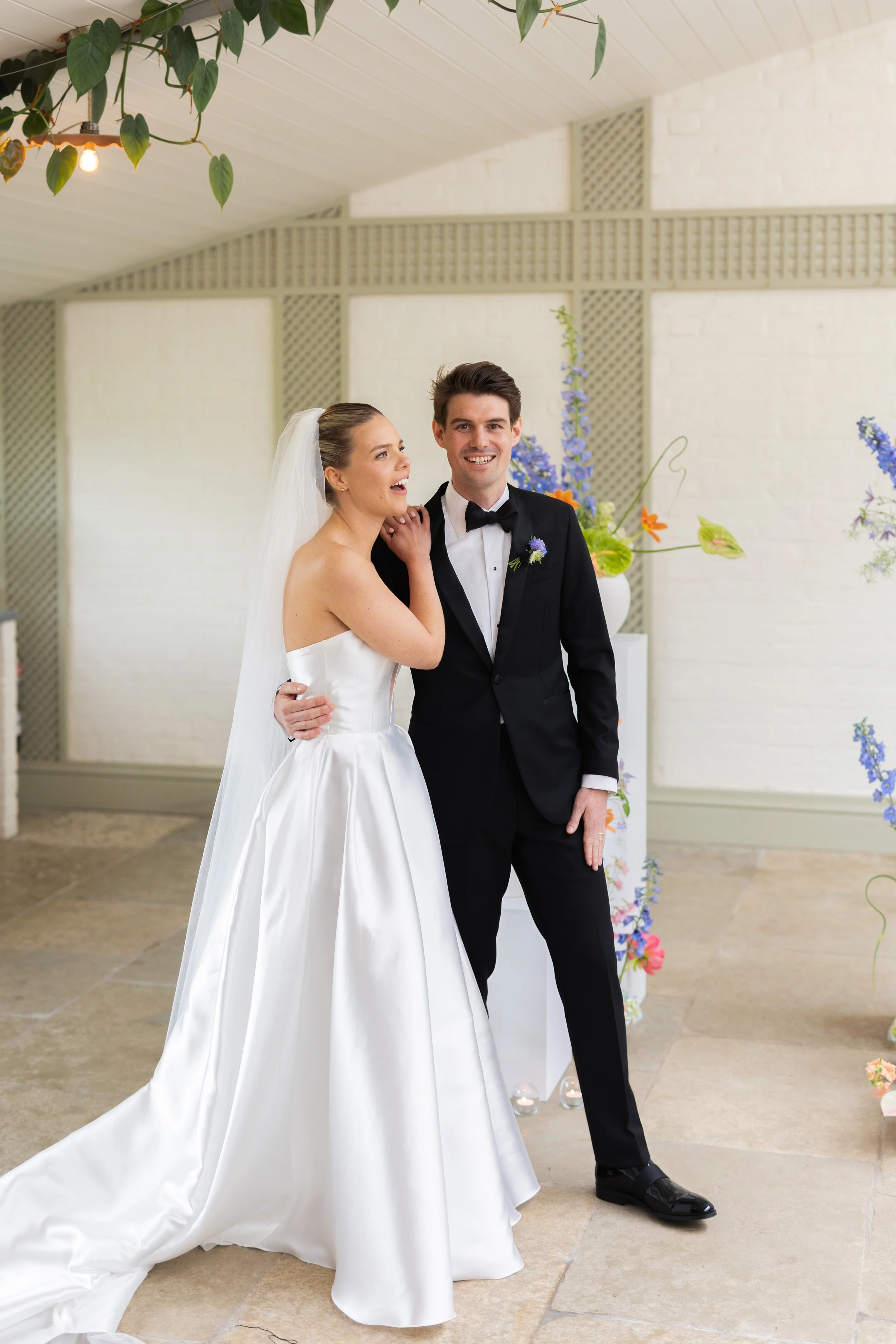 A bride and groom standing together indoors, smiling, with wedding attire, and floral decorations in the background.