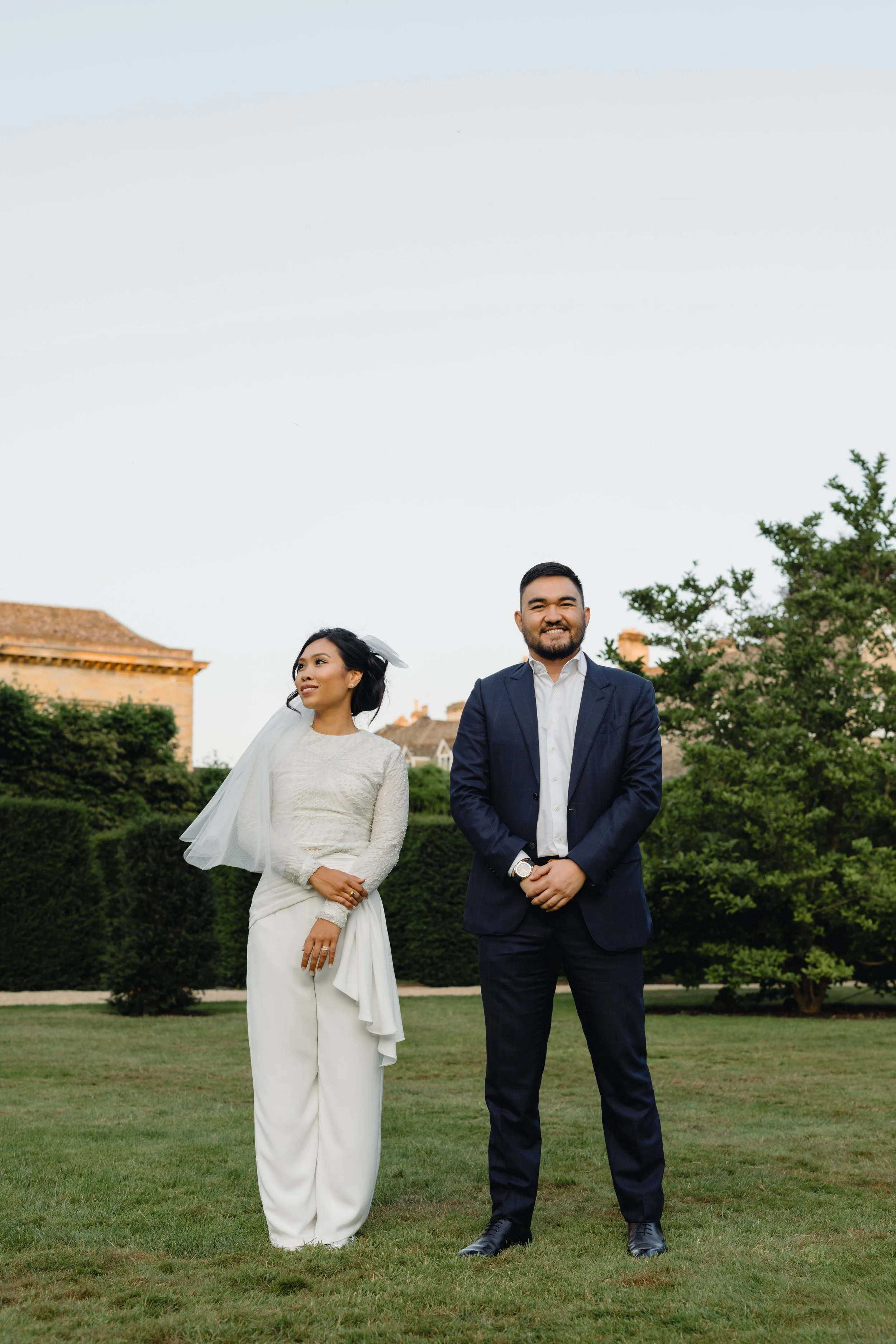 A bride and groom standing outdoors on a lawn, with the bride in a white wedding dress and veil, and the groom in a dark blue suit, smiling and holding hands.