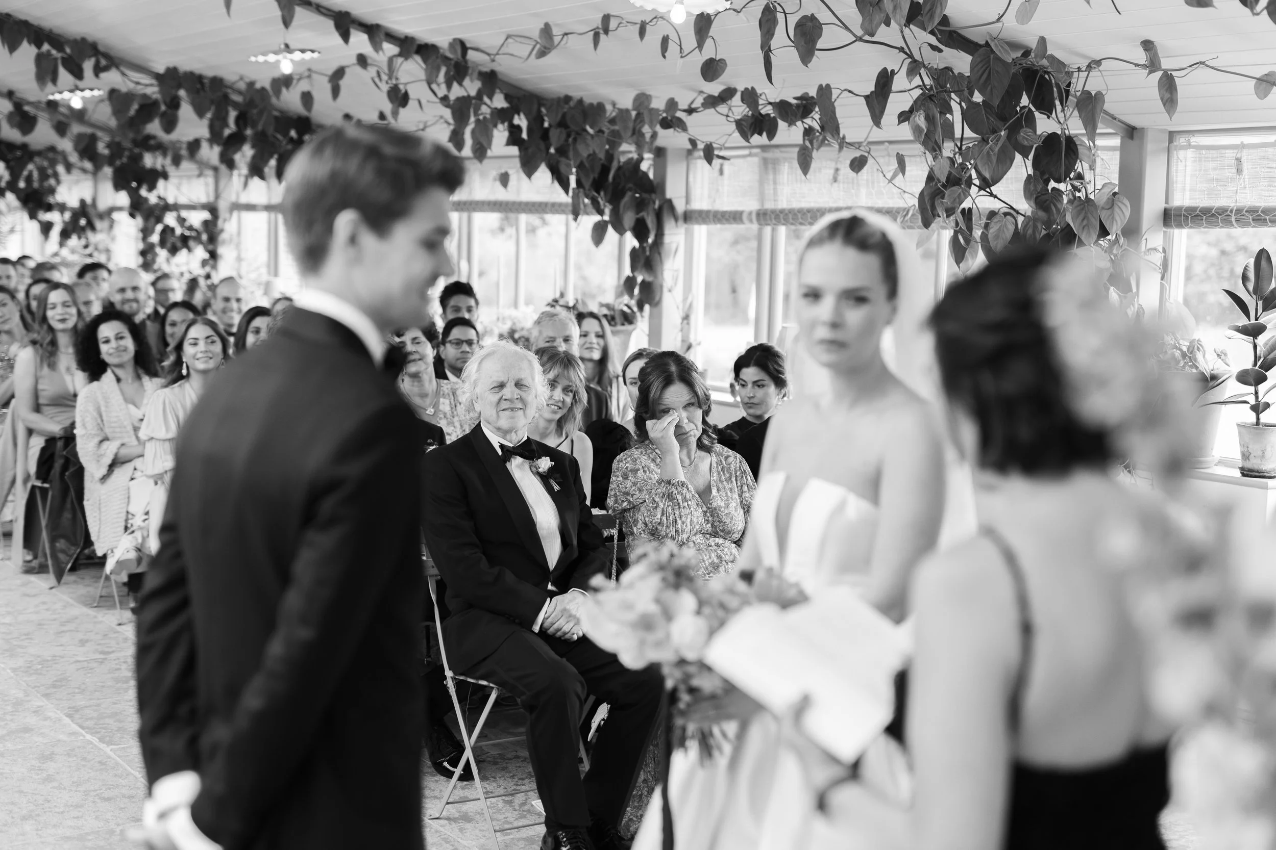 A wedding ceremony indoors with a groom and bride in focus, facing each other. Guests are seated in the background, some looking emotional, with plants and large windows visible.