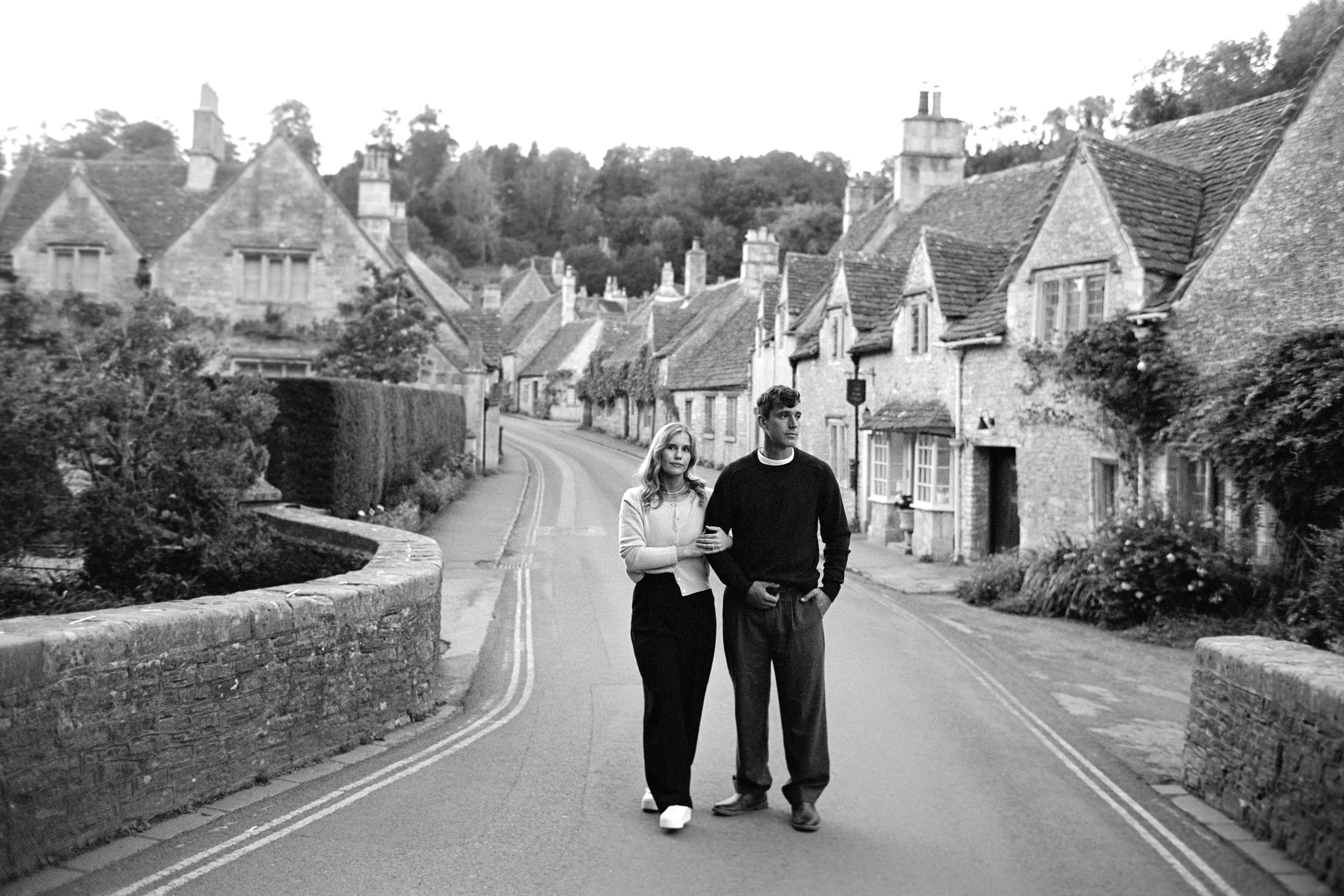 A black and white photo of a young couple walking arm in arm down a quaint, tree-lined street in a small village, with old stone houses and greenery on both sides.