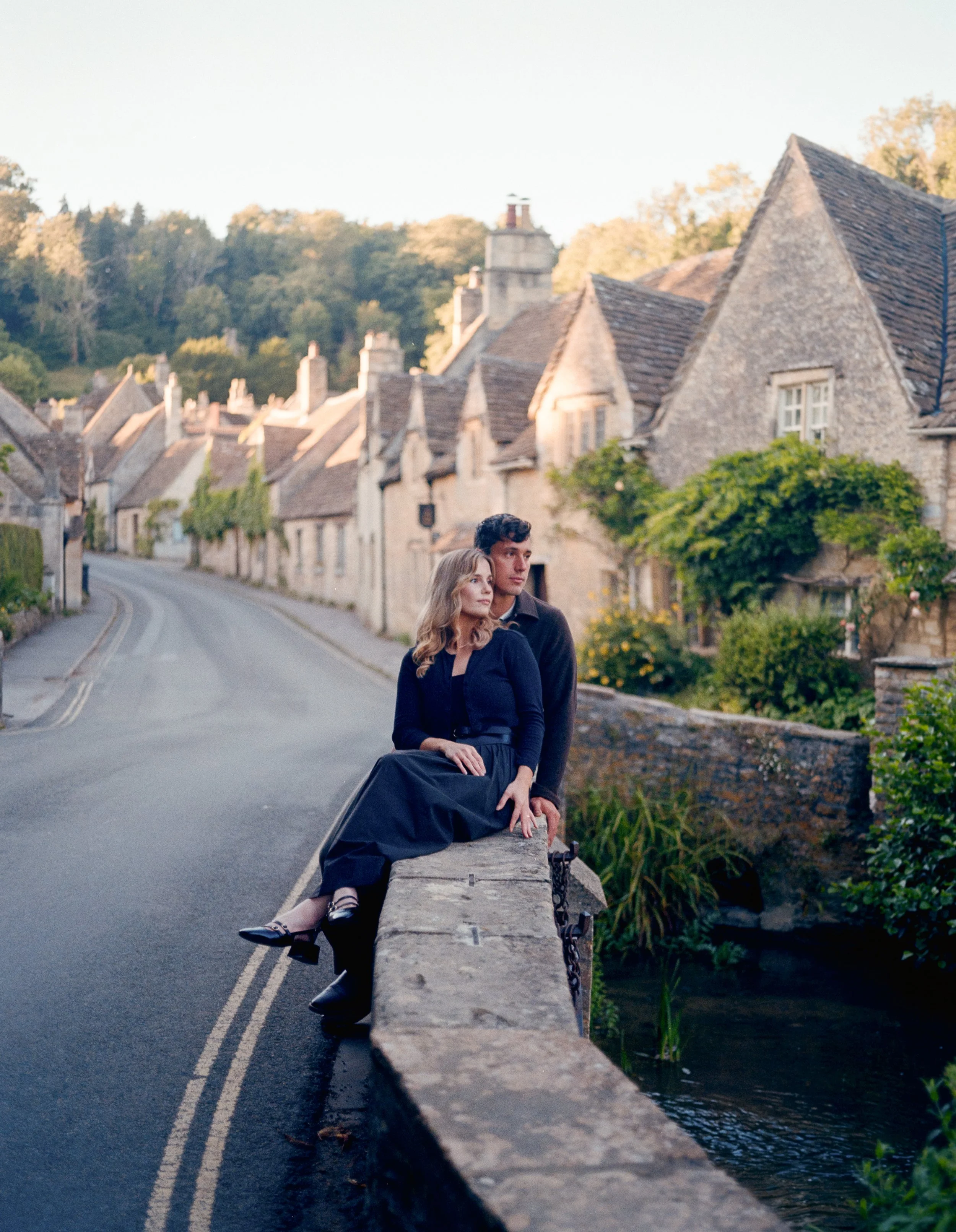A woman and a man sitting on a stone bridge over a small stream in a quaint village with stone houses and lush greenery.