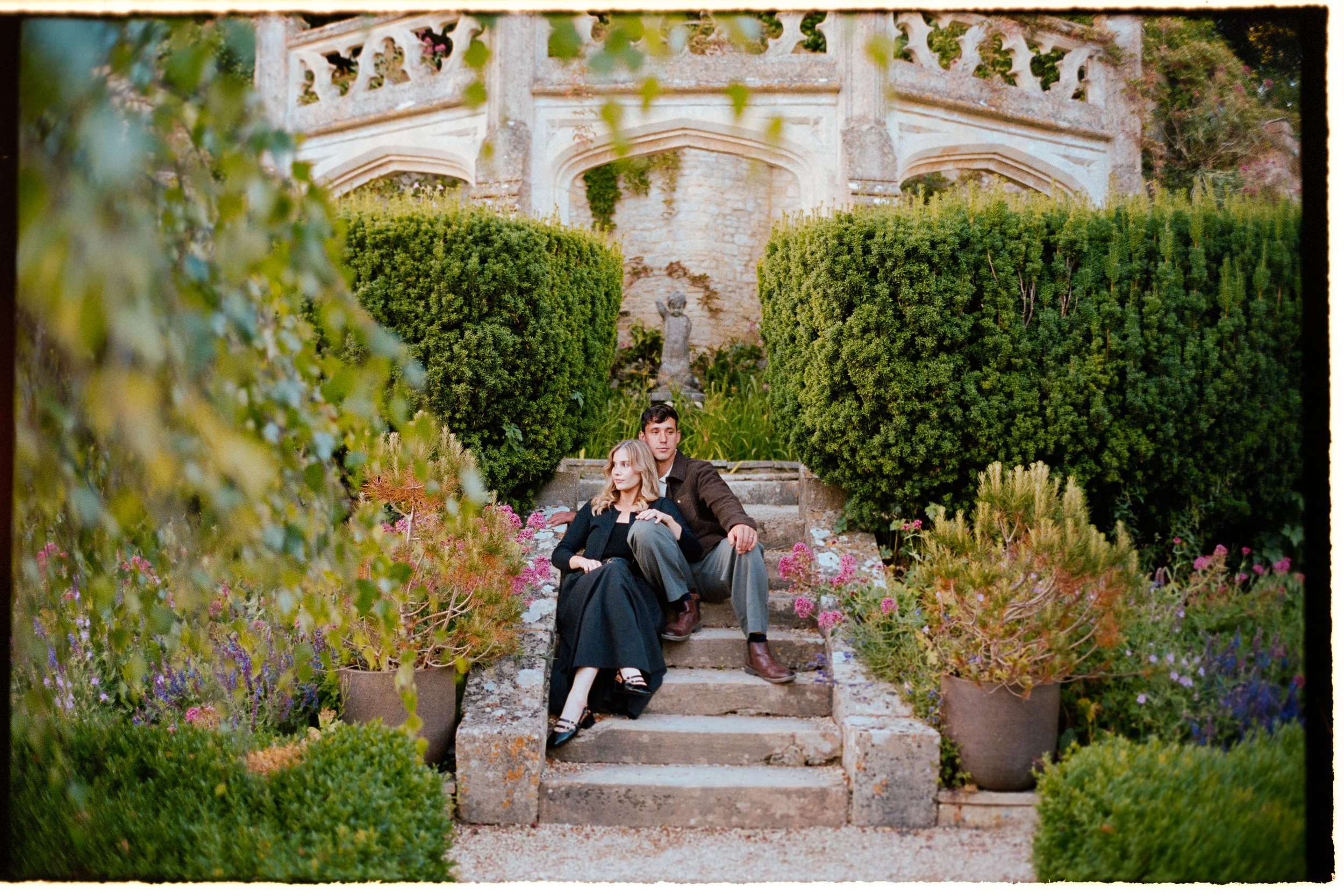 A young couple sitting on stone steps in a lush garden with large, trimmed bushes and colorful flowers, surrounded by classical architecture.