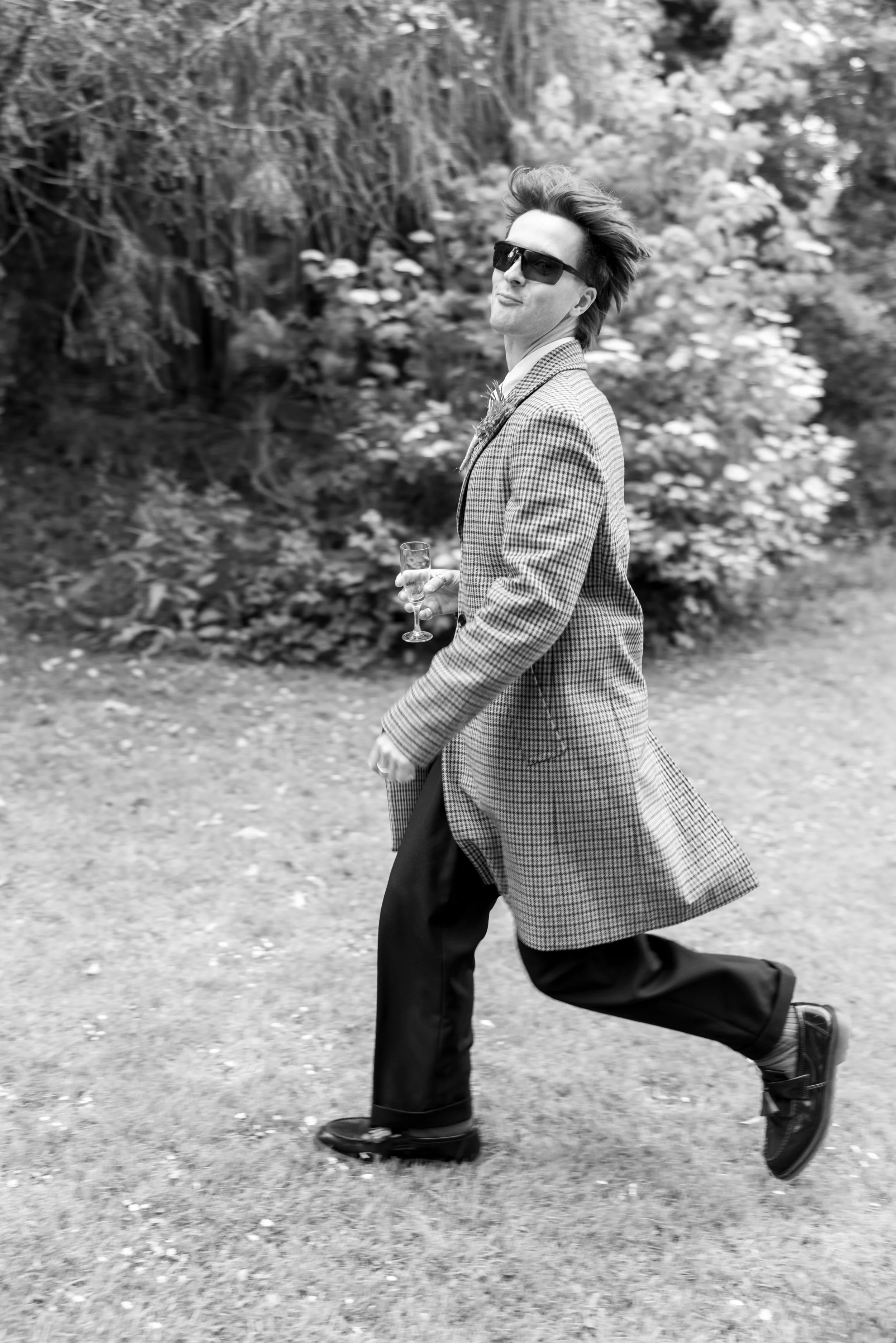 A man wearing sunglasses, a checkered blazer, black pants, and sneakers walks on grass while holding a champagne flute, with a blurred garden or park background.