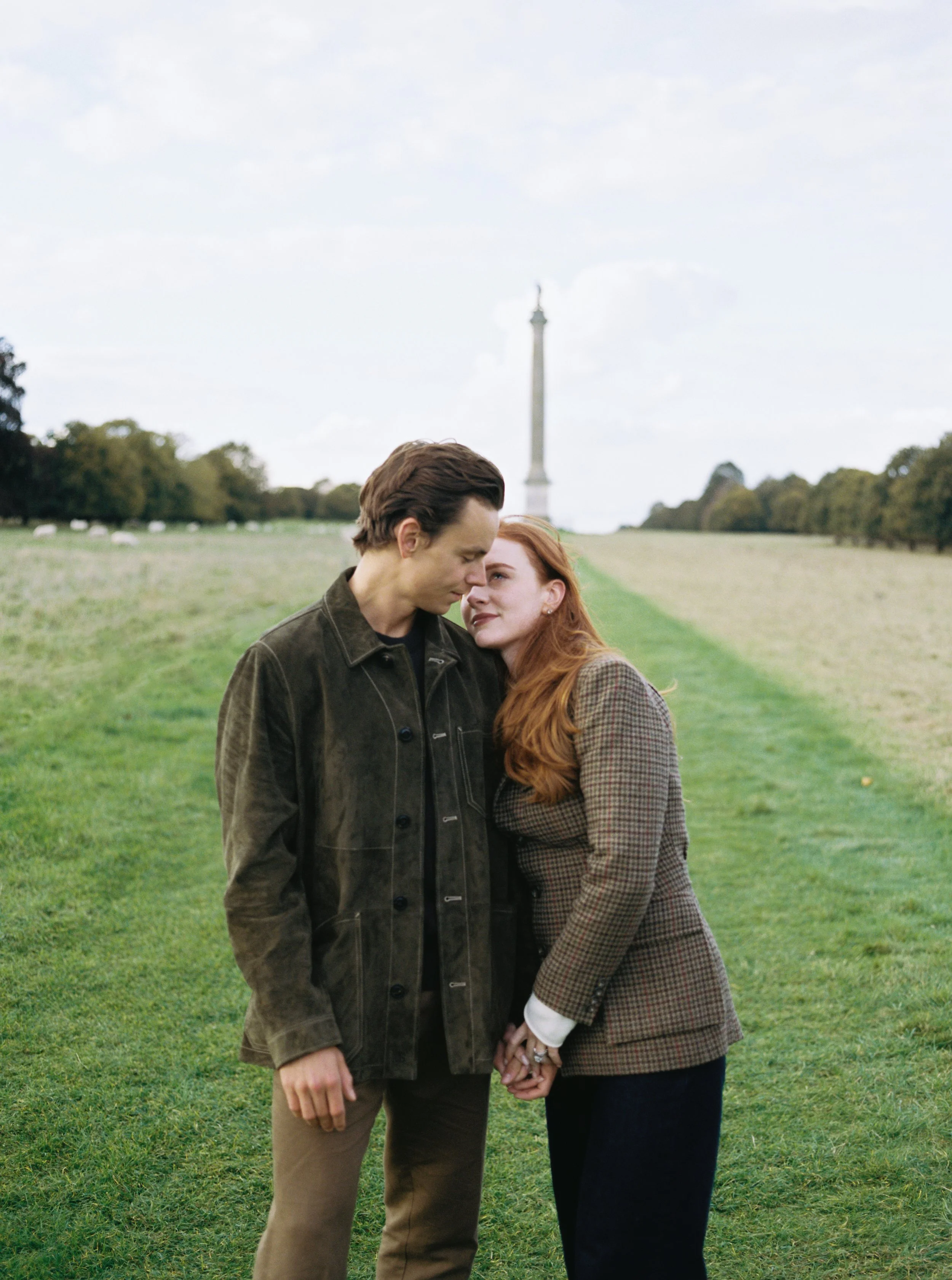 A couple holding hands and leaning their foreheads together outdoors on a grassy field, with a tall monument in the background and trees lining the distance.