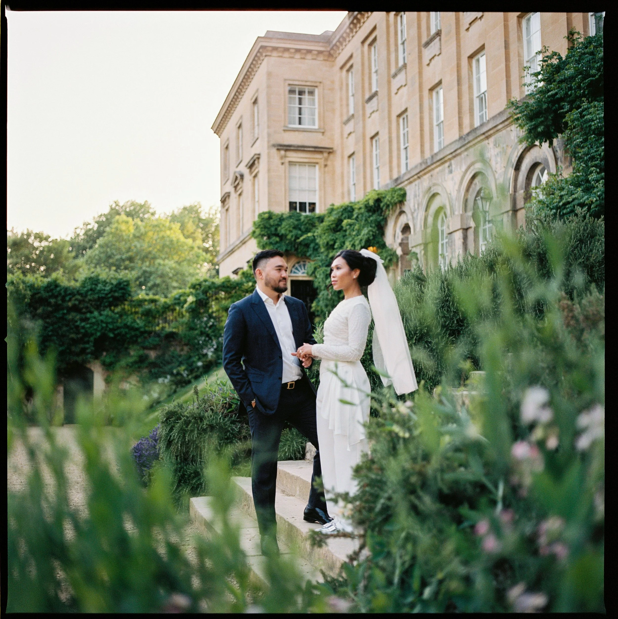 A bride and groom standing on a garden step in front of a historic building with ivy-covered walls, holding hands and looking at each other, surrounded by greenery and flowers in outdoor wedding attire.