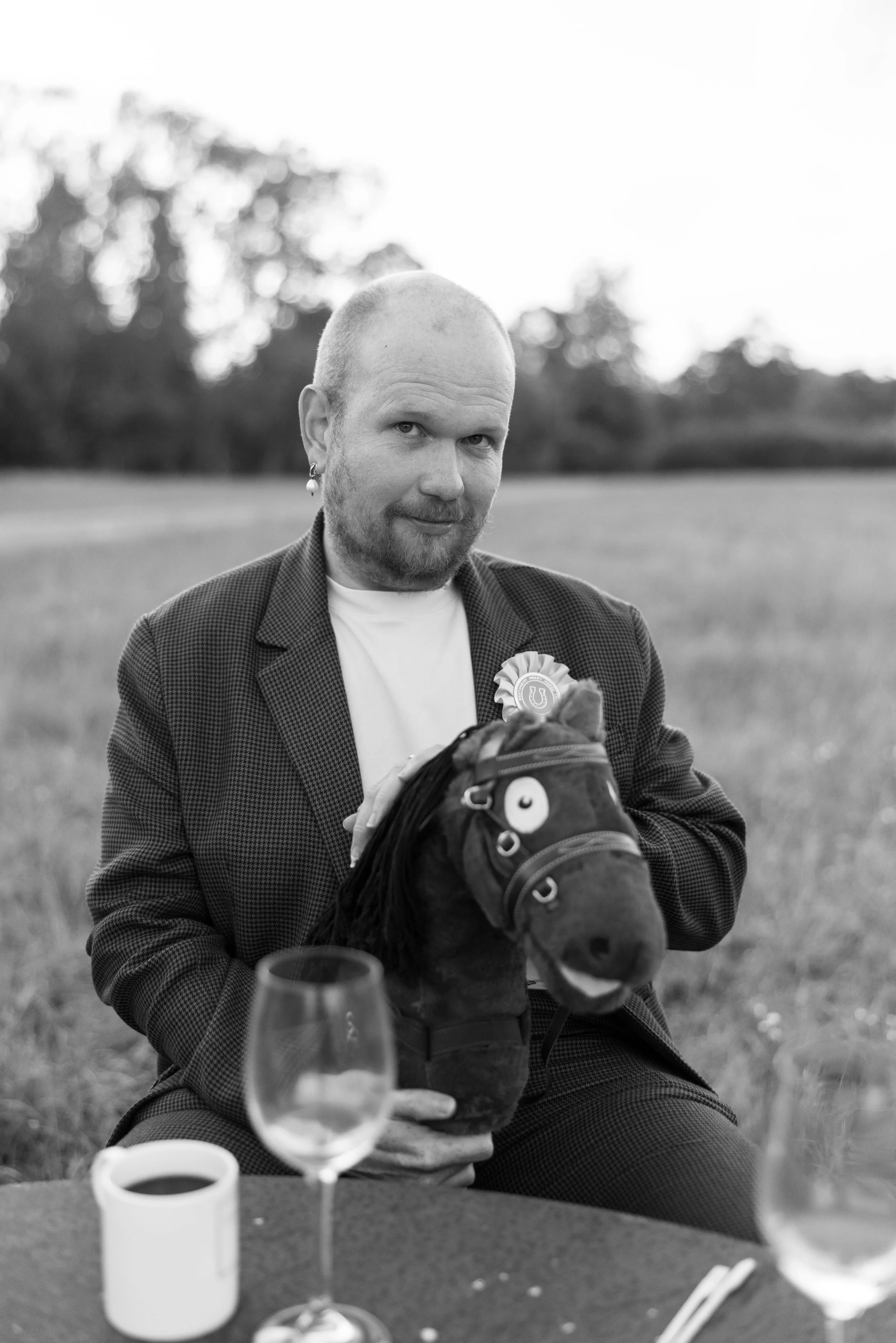 A man with a beard and a bald head, wearing a blazer, sitting outdoors holding a toy horse head, with a picnic table in front and wine glasses nearby.