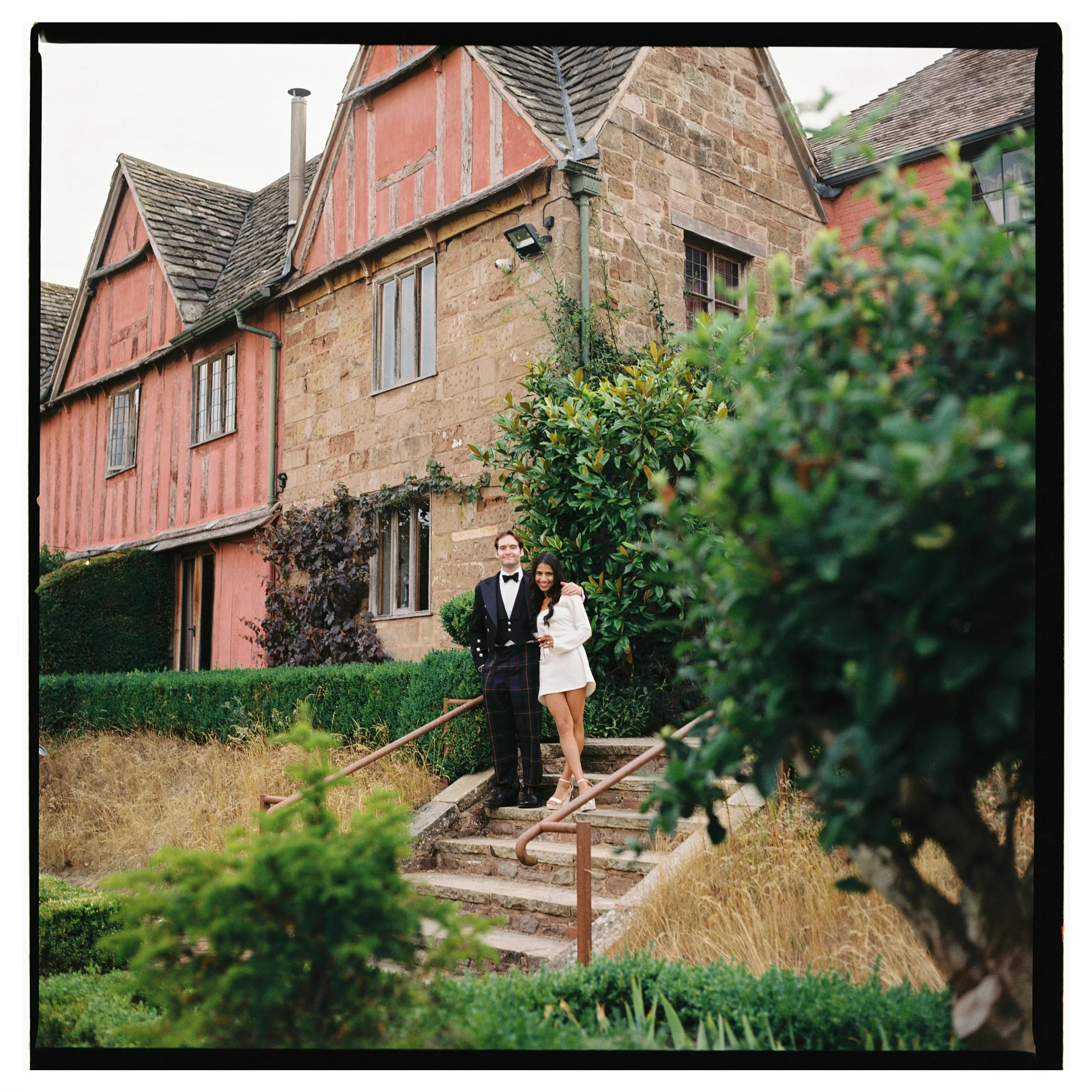 A bride and groom dressed in formal attire standing on outdoor stone steps in front of historic wedding venue Pauntley Court.