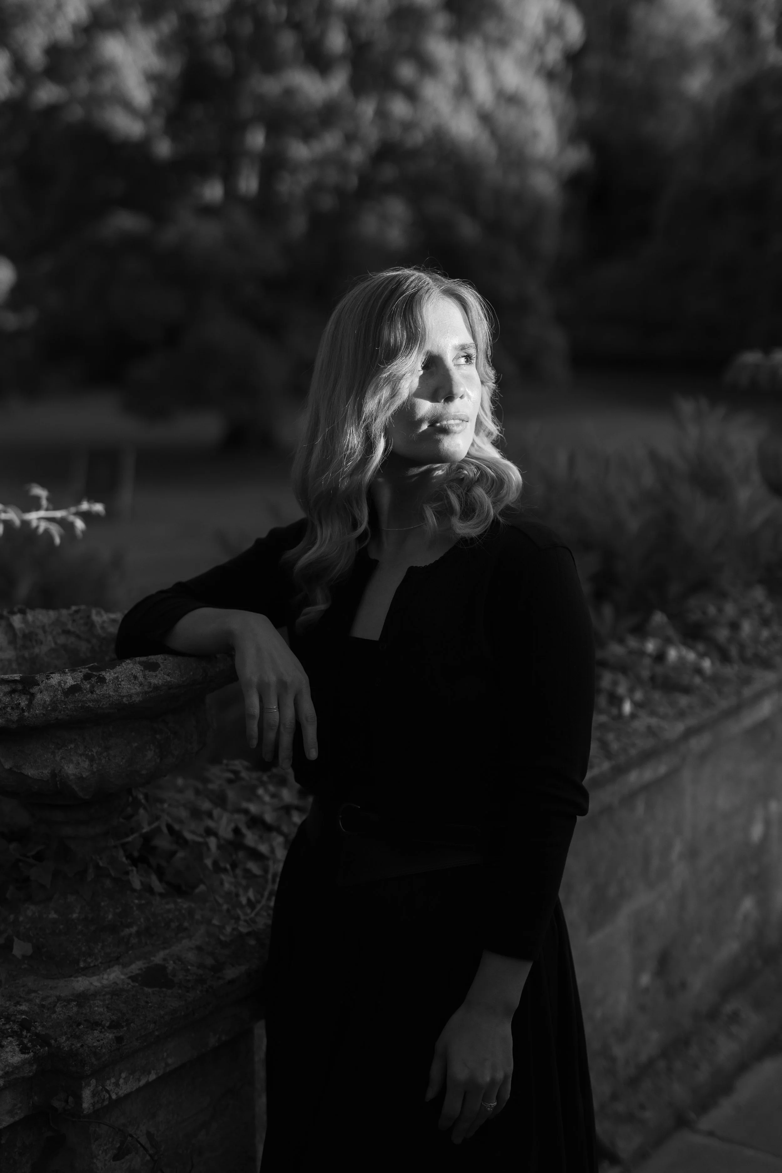 A woman with wavy hair stands outdoors in the evening, gazing into the distance, illuminated by the setting sun.