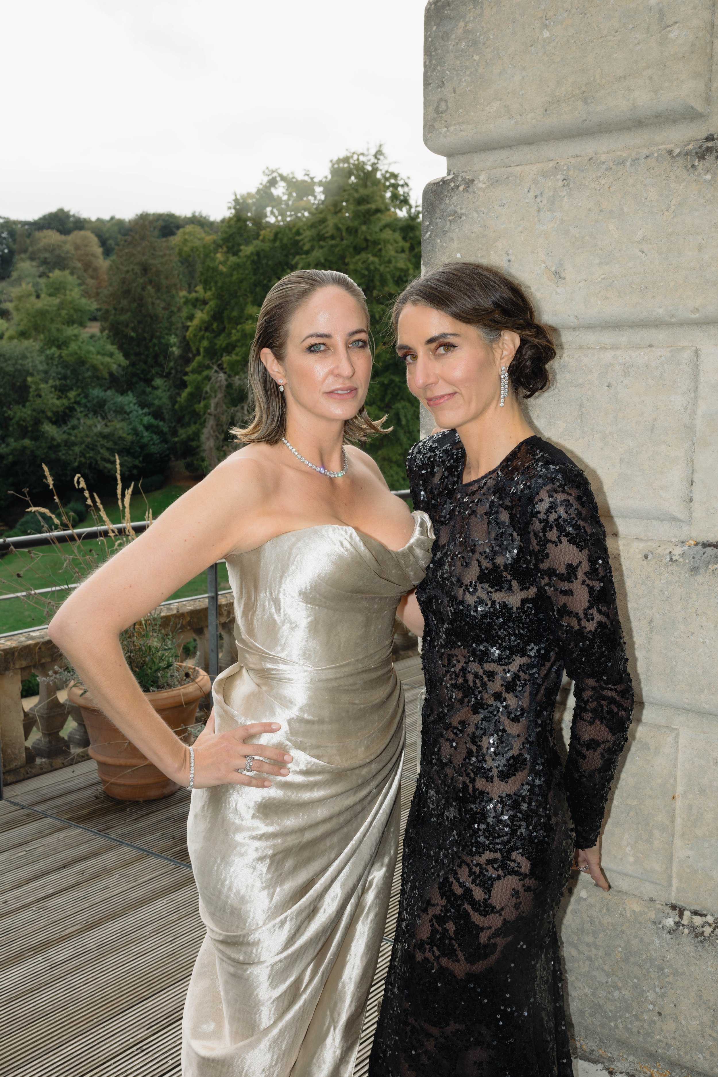 Two women in formal dresses standing against a stone pillar outdoors, with greenery and cloudy sky in the background.
