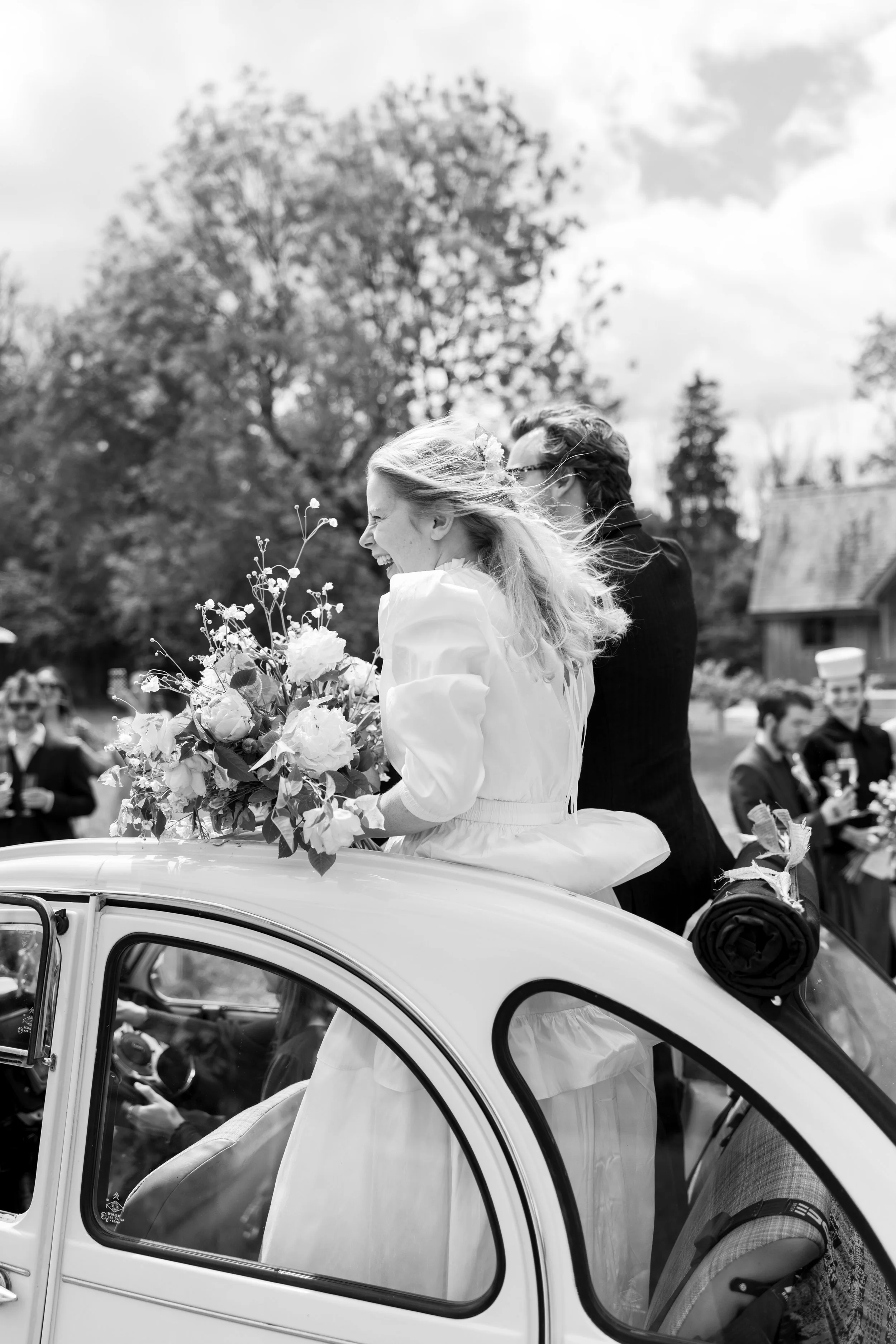 Black and white photo of a bride and groom riding in a vintage car during a wedding celebration, with guests in the background.