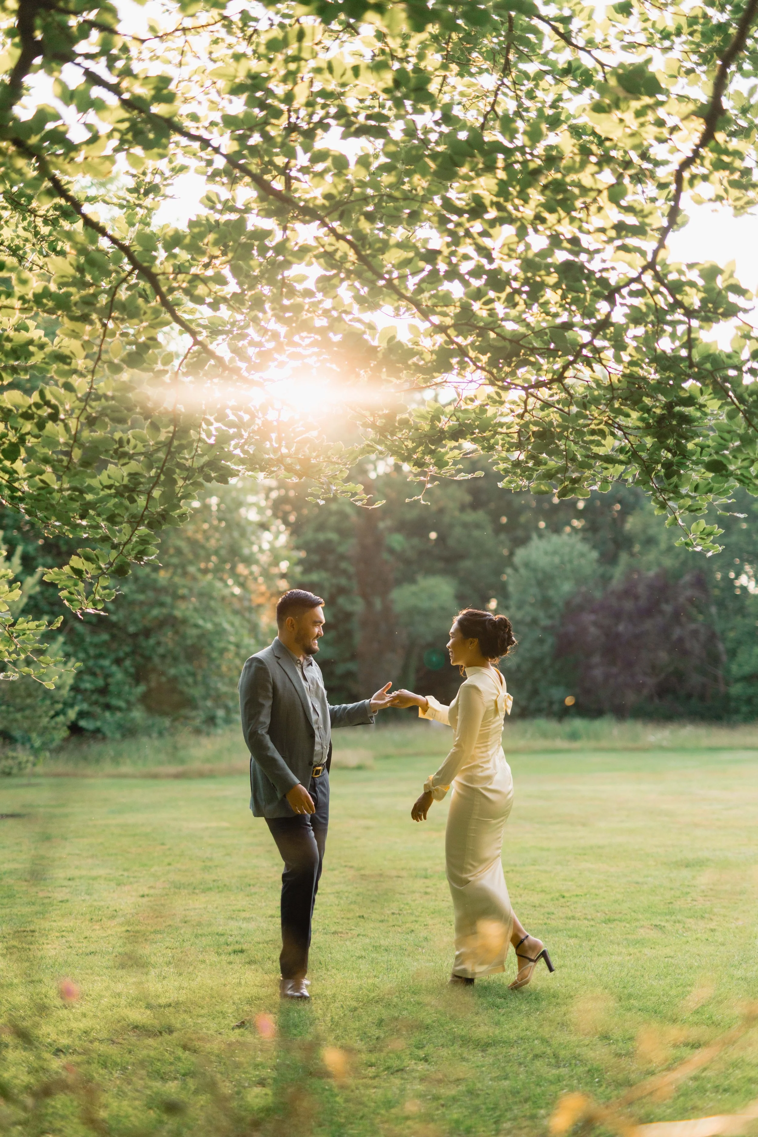 A man and woman are dancing outdoors in a park during sunset, holding hands and smiling at each other.