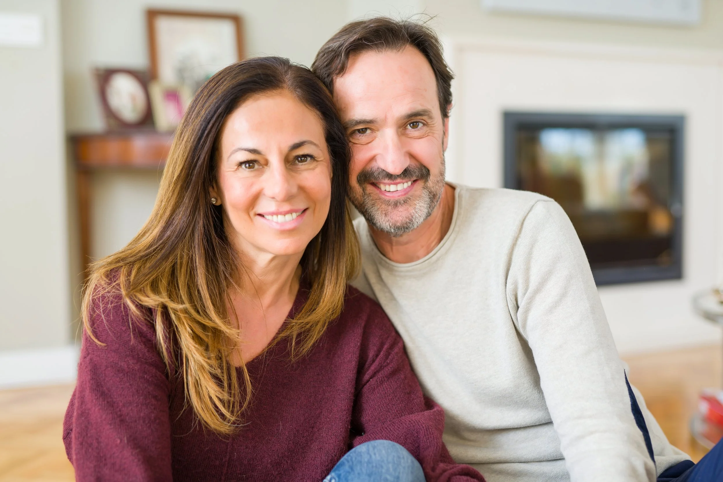 A smiling couple, a woman with long brown hair and a man with dark hair and a beard, sitting close together indoors in front of a TV.