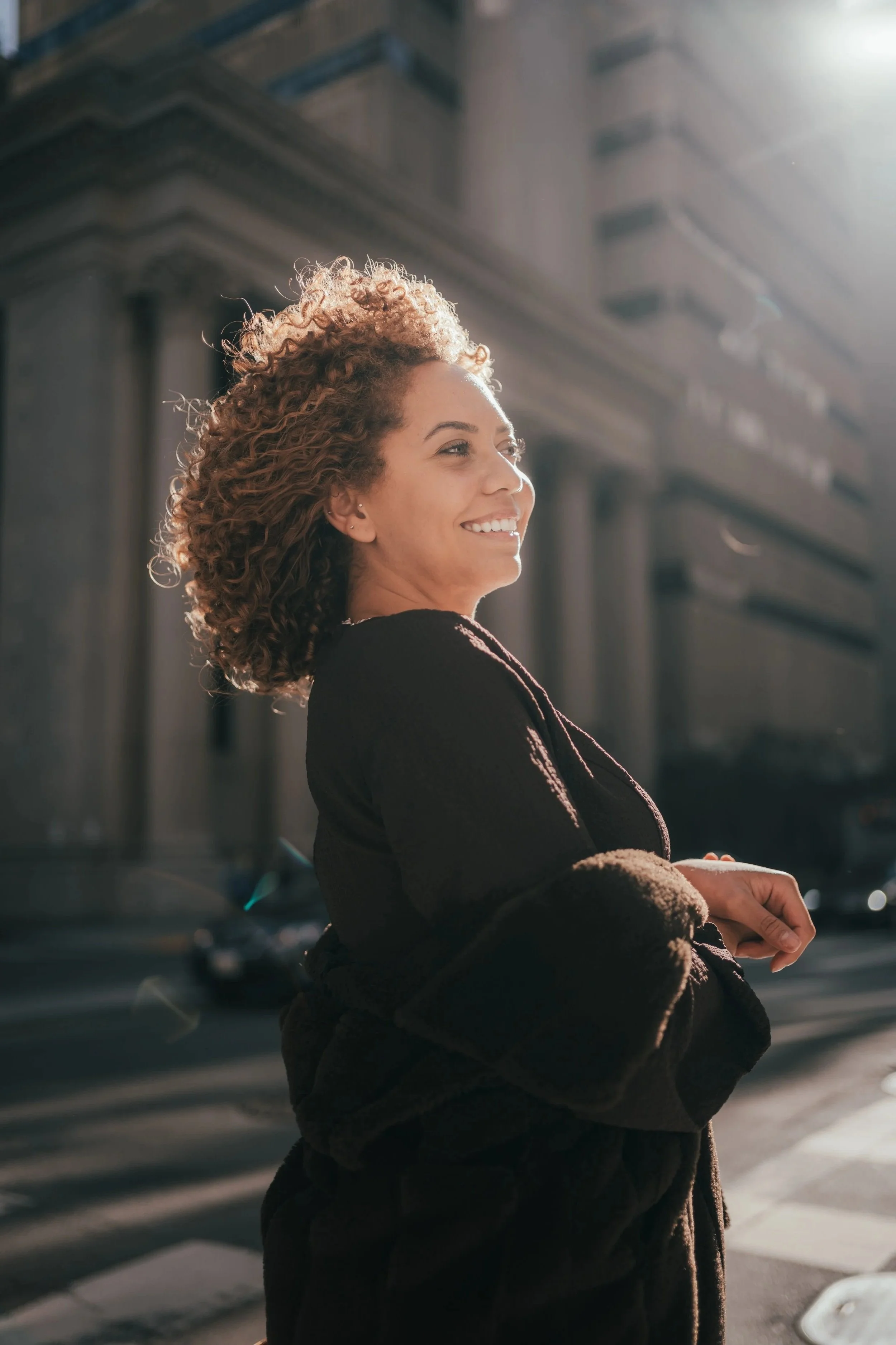 A woman with curly hair smiling, standing outdoors in sunlight, with a city street background.