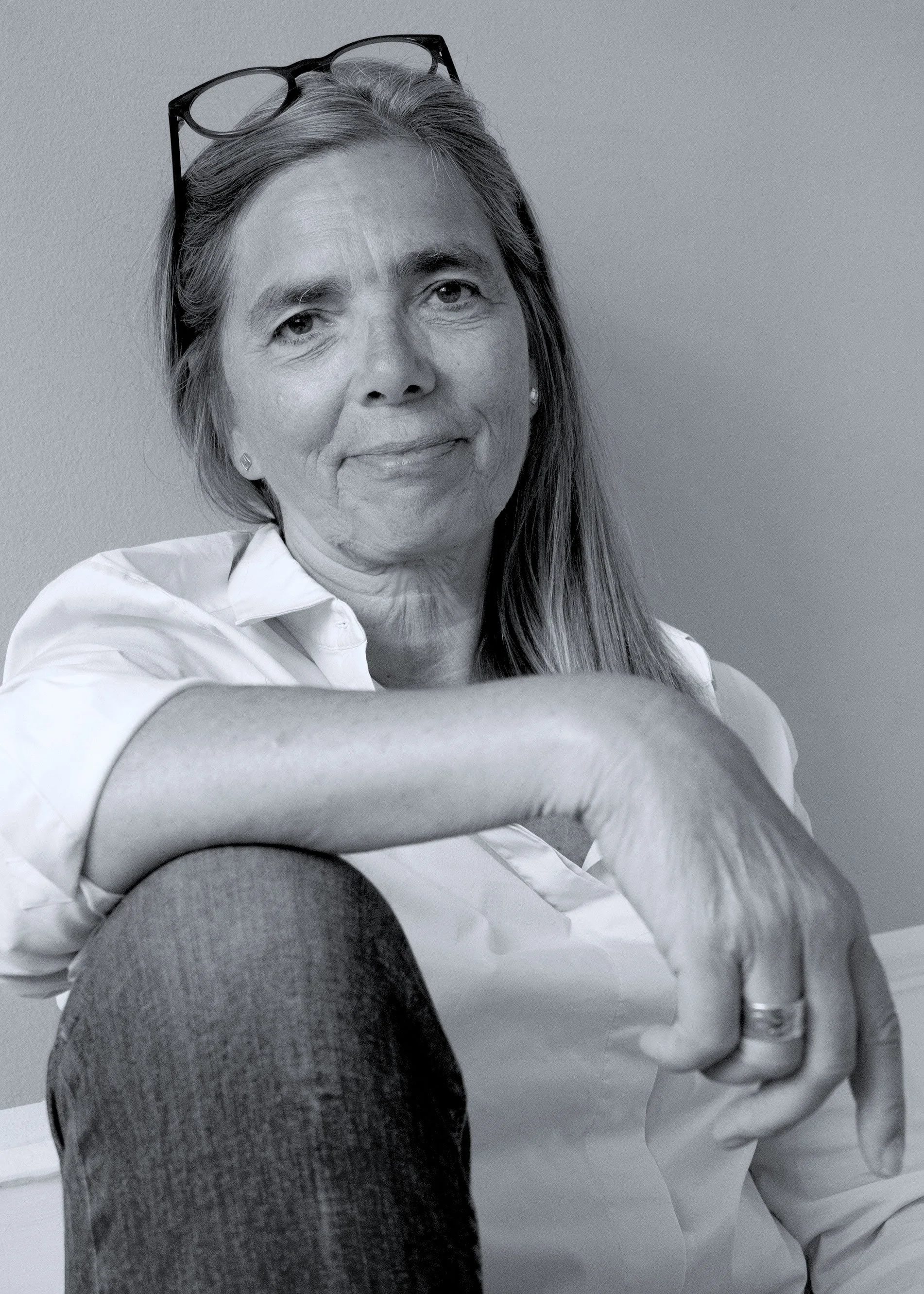 Black and white portrait of Susanne Grønlund with glasses on her head, wearing a white shirt and jeans, seated with her arm resting on her knee.