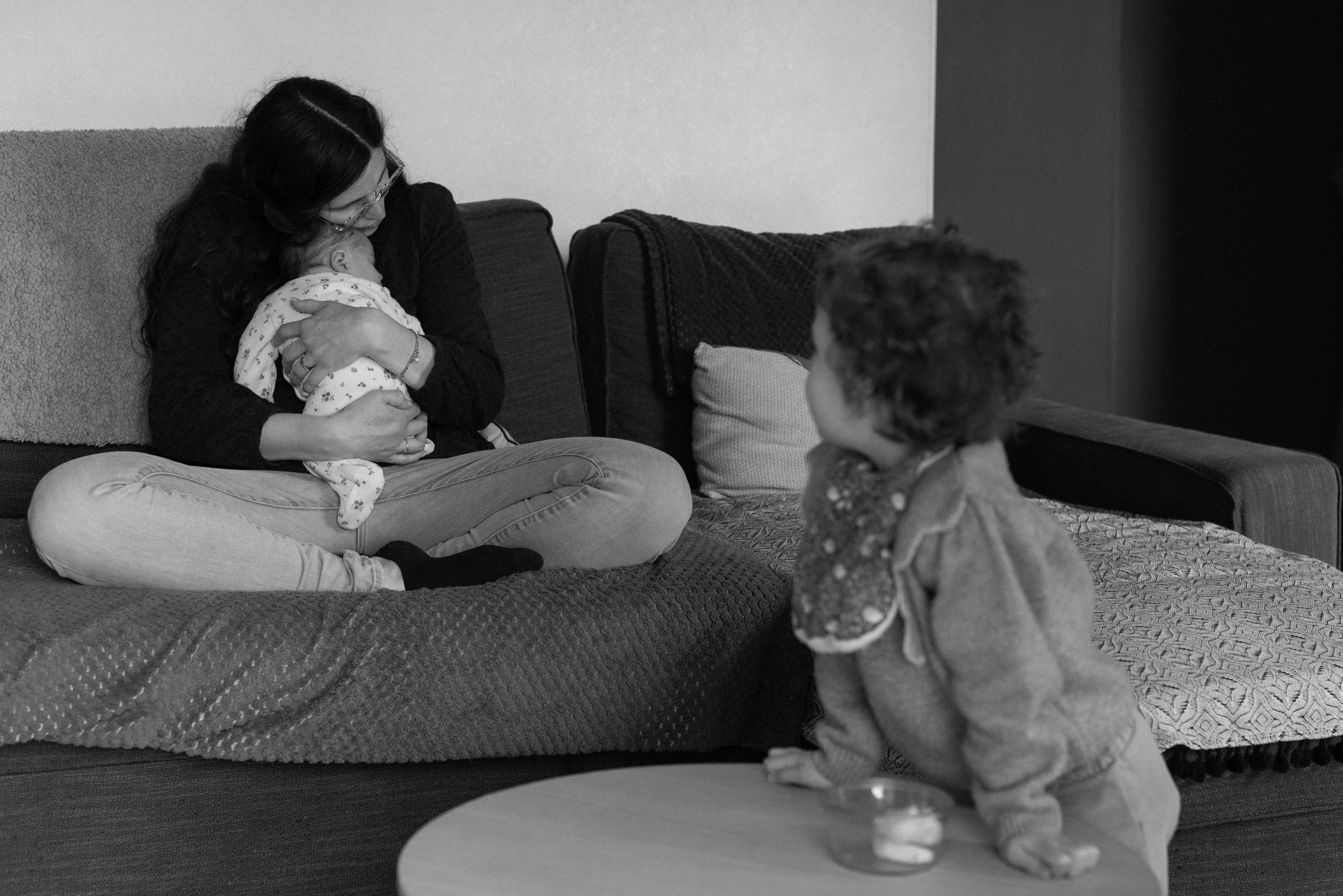 une maman sur le canapé en calin avec son bébé et la grande sœur les regardant au premier plan