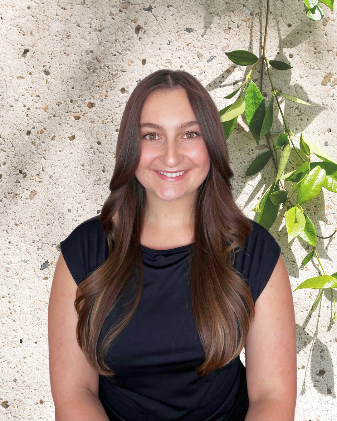 A woman with long brown hair wearing a black top, smiling, standing in front of a textured wall with a green leafy plant hanging behind her.
