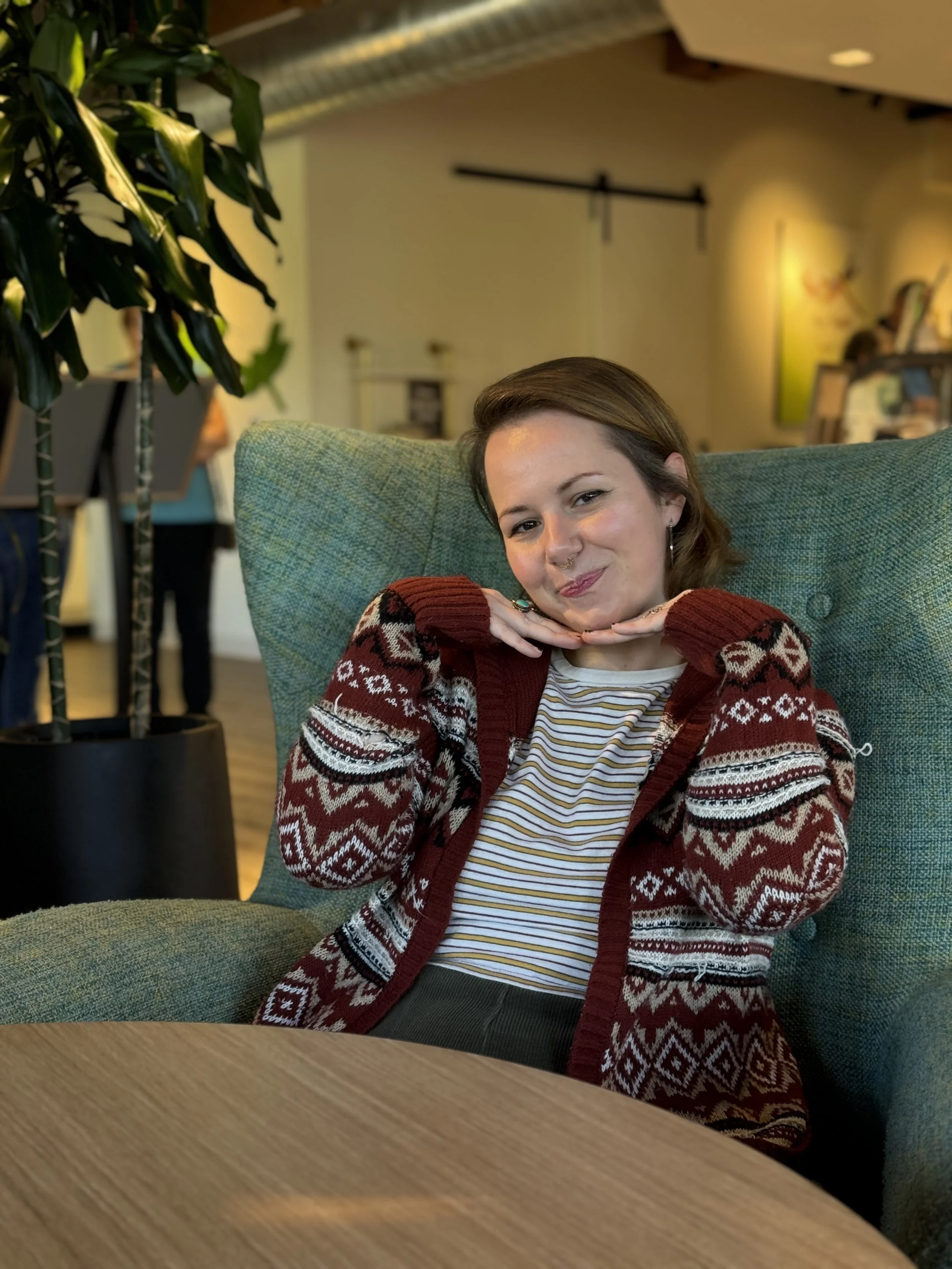 A woman with short brown hair and a septum piercing sitting in a green armchair, smiling and posing with her hands under her chin. She is wearing a striped shirt and a red, white, and black patterned sweater. The background shows a modern, cozy interior with artwork on the walls and a large potted plant to her left.