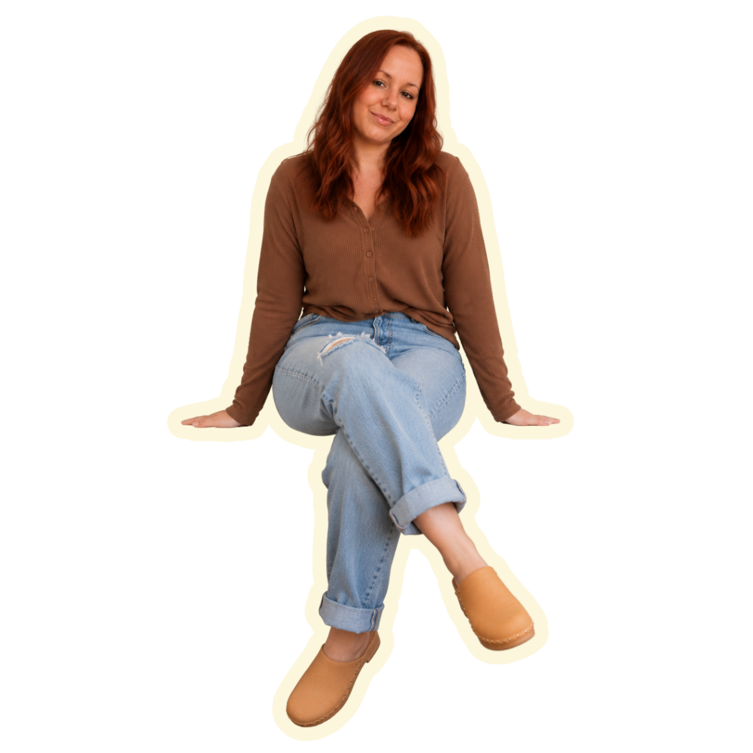 A woman with red hair sitting on the ground, wearing a brown cardigan, light blue ripped jeans, and tan boots, smiling and looking at the camera. She is posing with one leg crossed over the other and her hands resting on the ground.