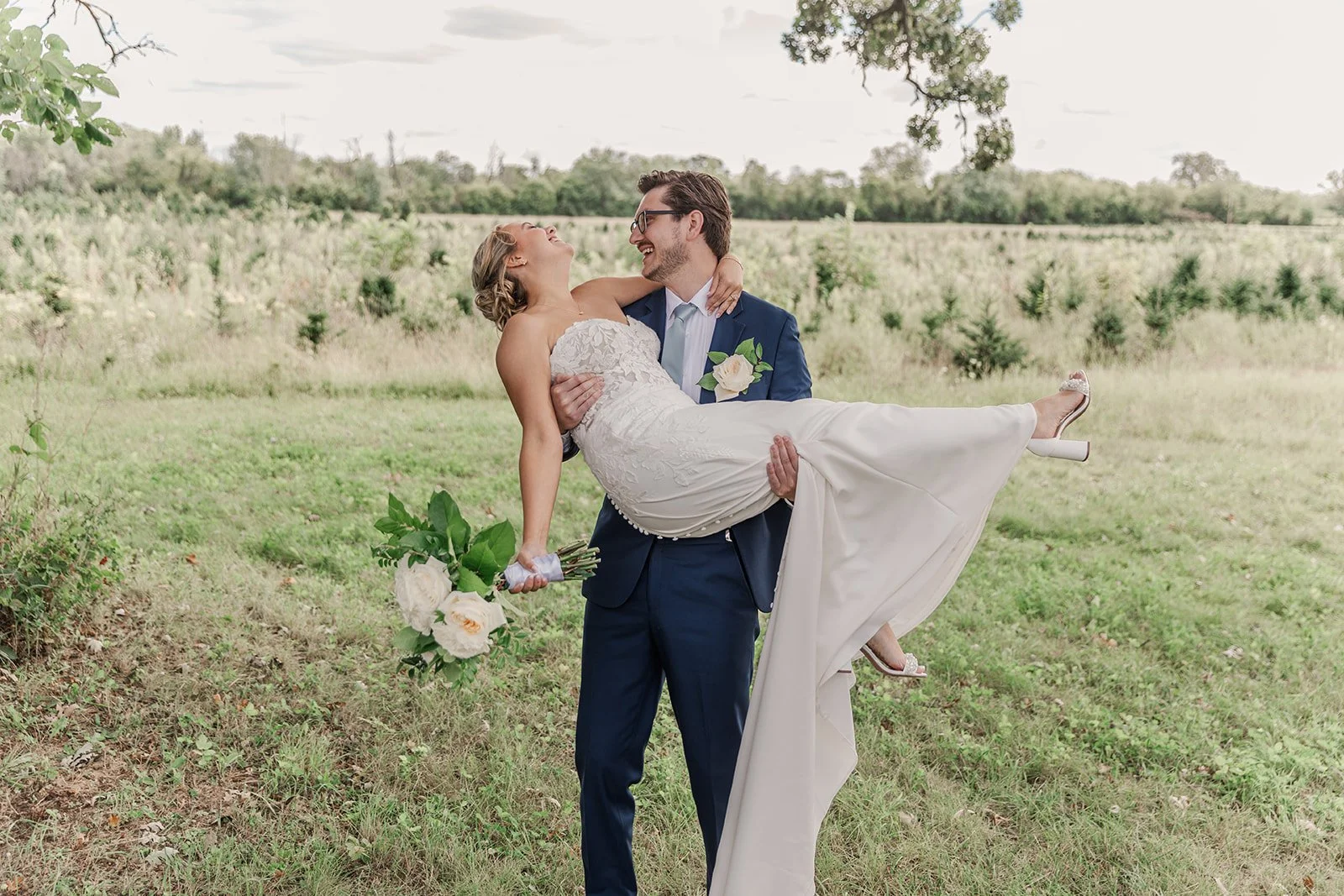 A bride in a white wedding dress being carried by a groom in a navy blue suit in a grassy outdoor setting, smiling and holding a bouquet of white roses.